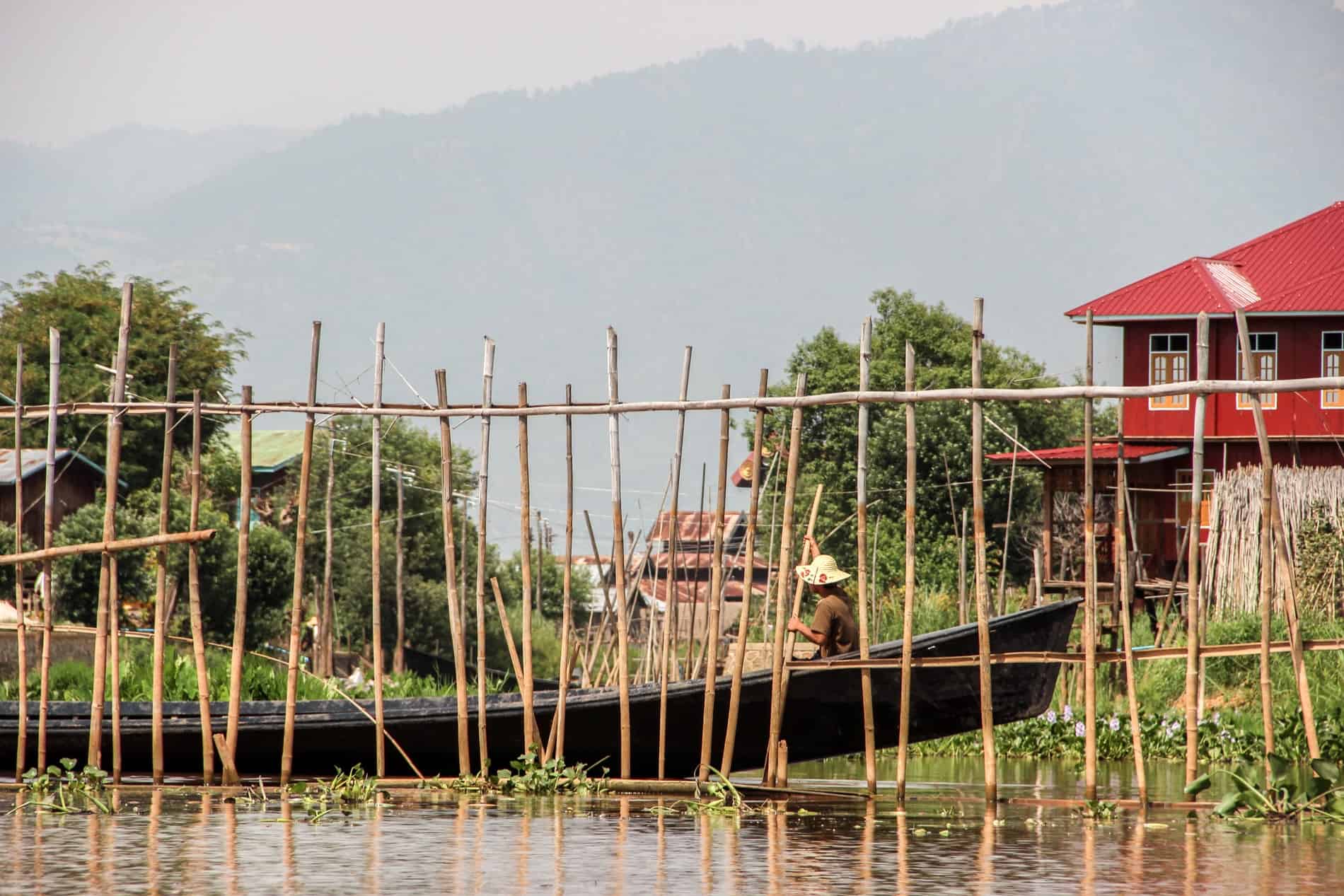 A person rows a long, dark wooden boat between a red houses and a bamboo pole wall in a floating garden on Inle Lake. 
