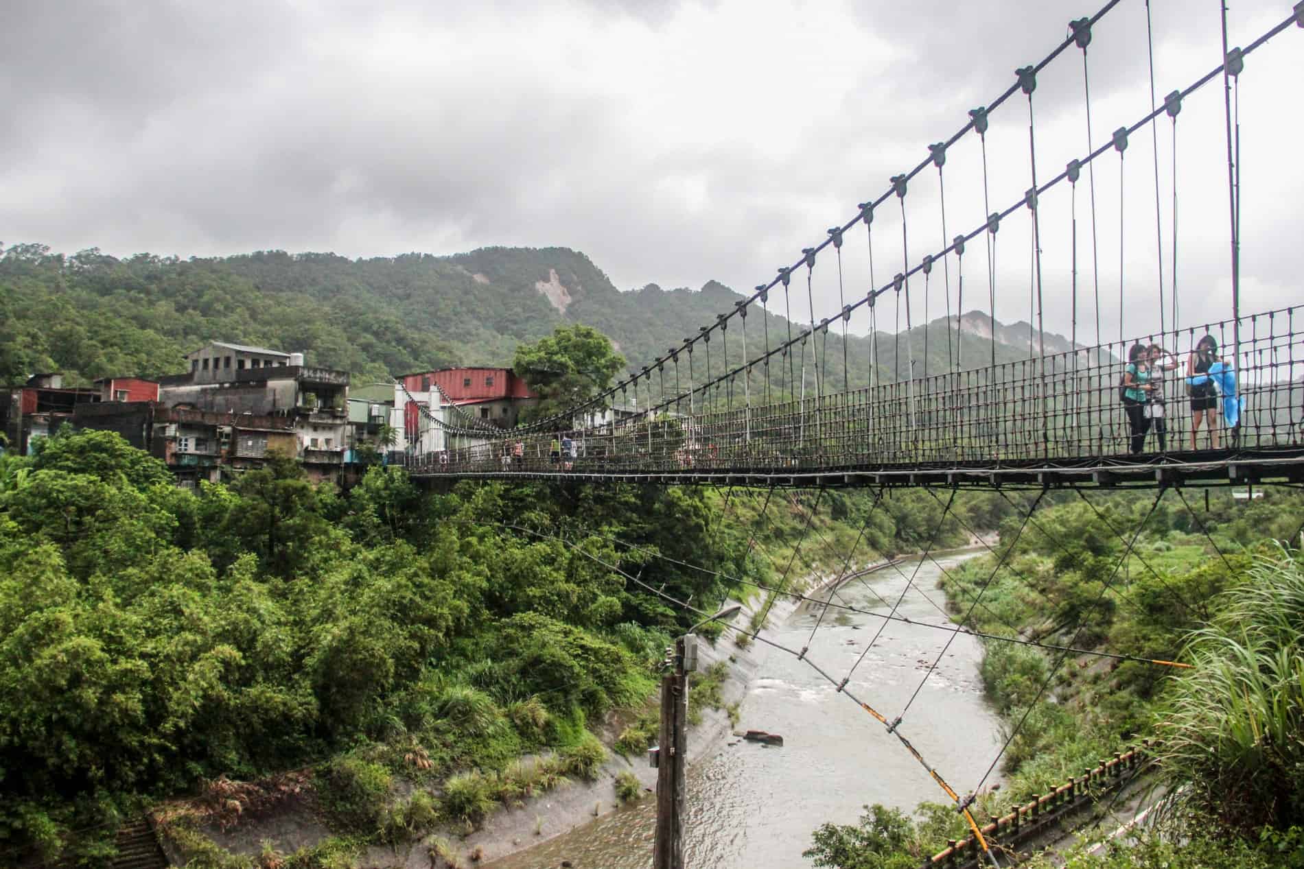 A small group of people at the end of a suspension bridge reaching across a river and connecting to a forested hilltop town. 