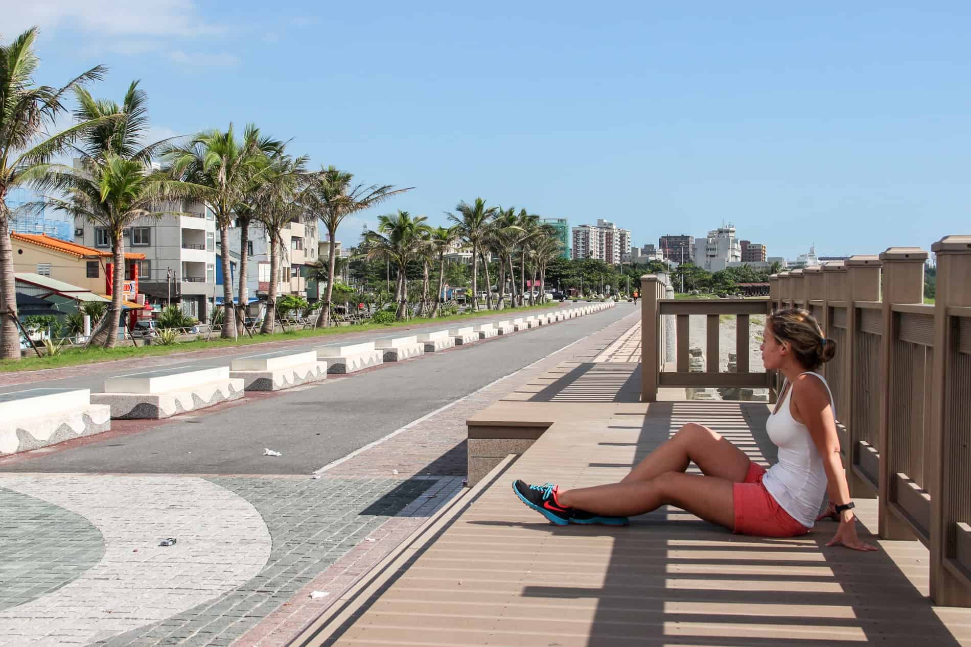 A woman in a white top and bright pink shorts sitting on a wooden jetty looking towards a tree-lined street with low-rise buildings. 