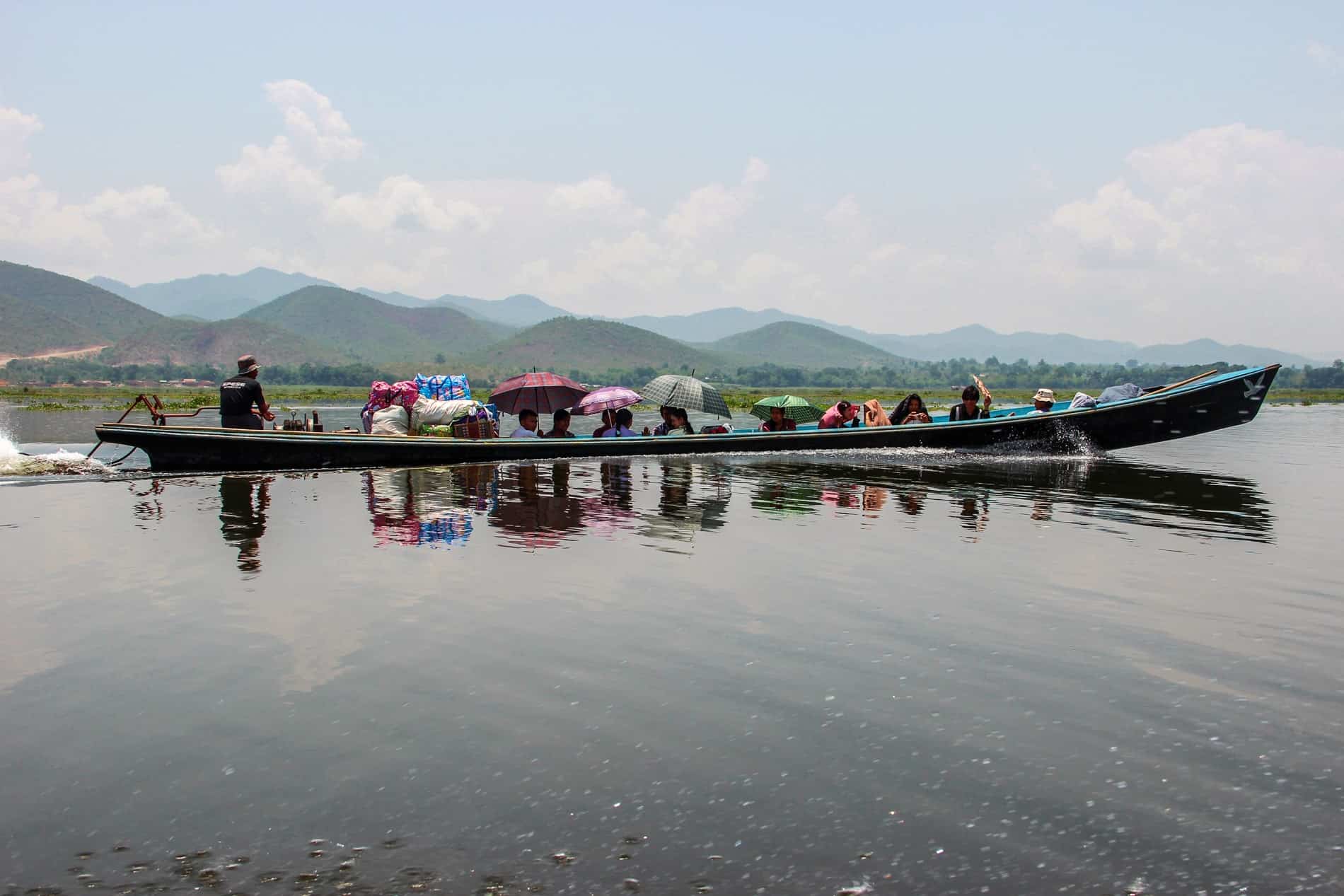 A line of people holding colourful umbrellas in a longboat, being transported across the waters of a large reservoir.
