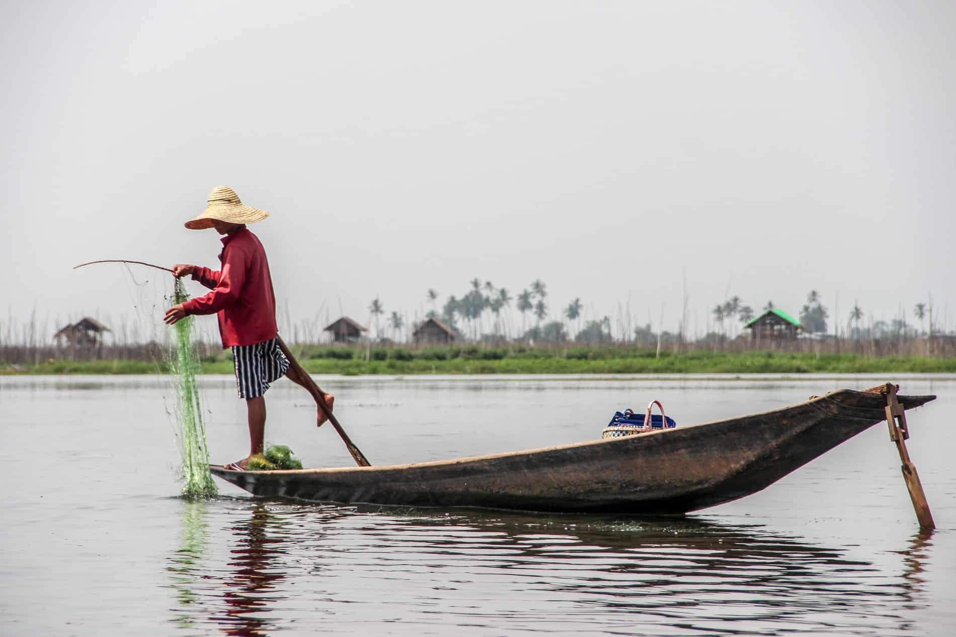 A fisherman in a red top, stripy shorts and a straw hat on Inle Lake in Myanmar practicing the traditional method of rowing the oar with one leg.