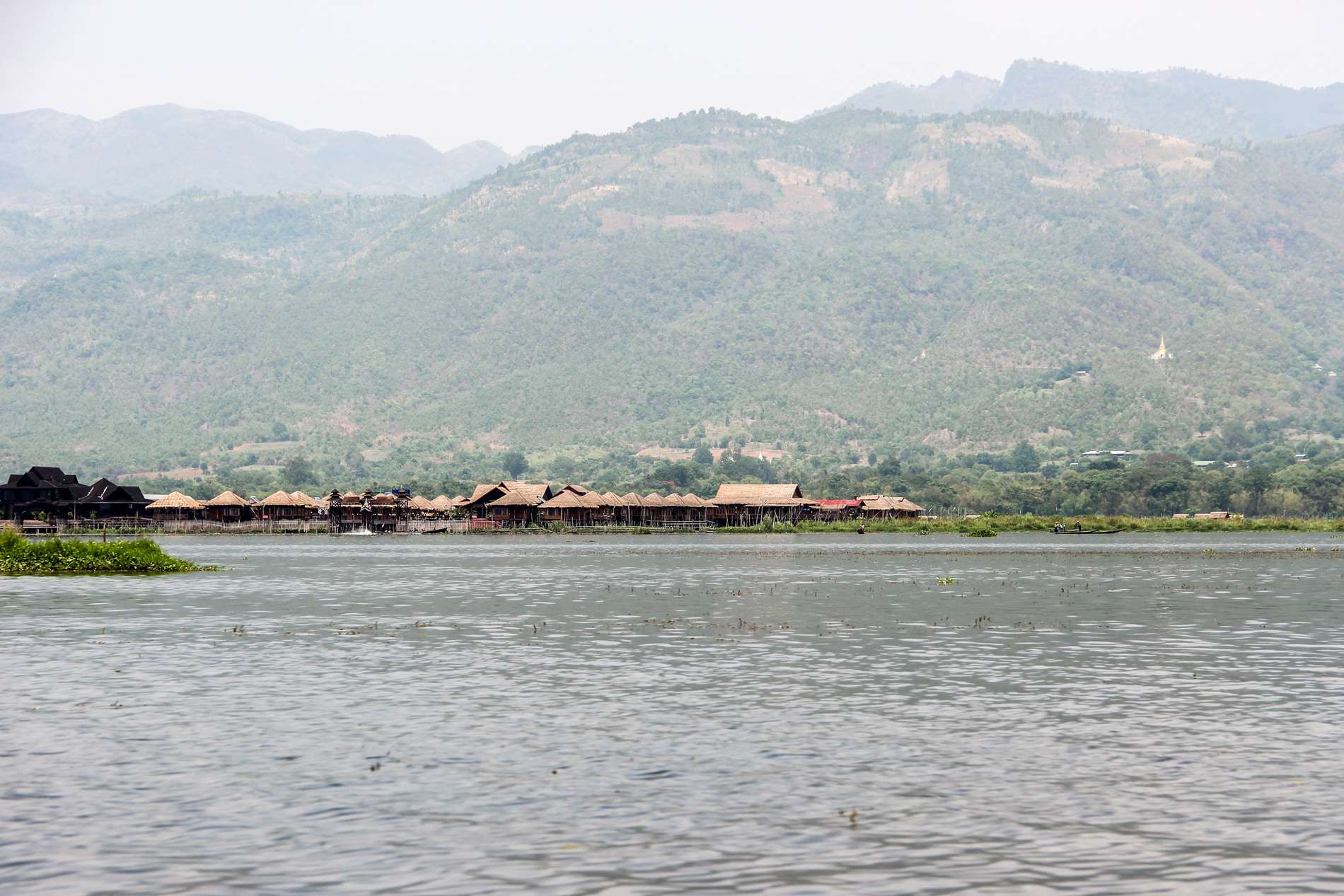 Distanced view of a community of bamboo stilt houses in the middle of the vast Inle Lake basin surrounded by mountains. 