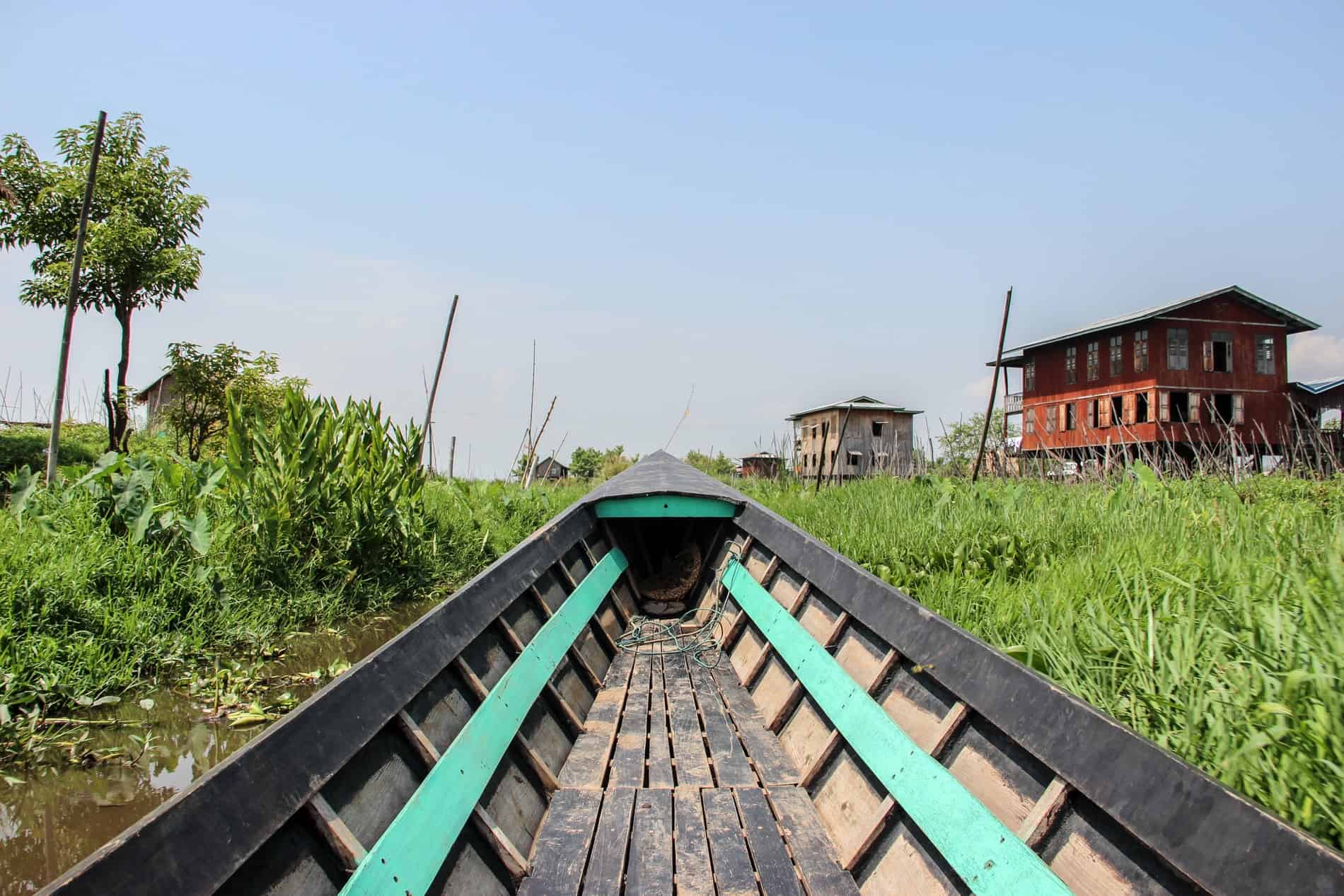 A long wooden boat with mint green paint colouring moves forward through a narrow canal on Inle Lake lined with floating green gardens and large wooden stilt houses. 