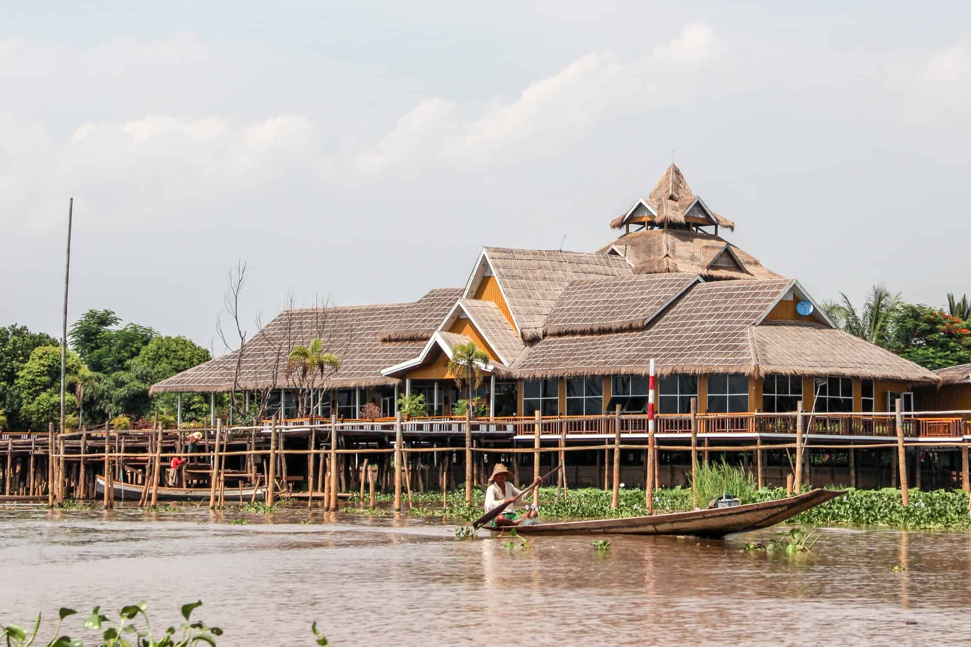 A man rows a long canoe past a huge lake side house on stilts. 