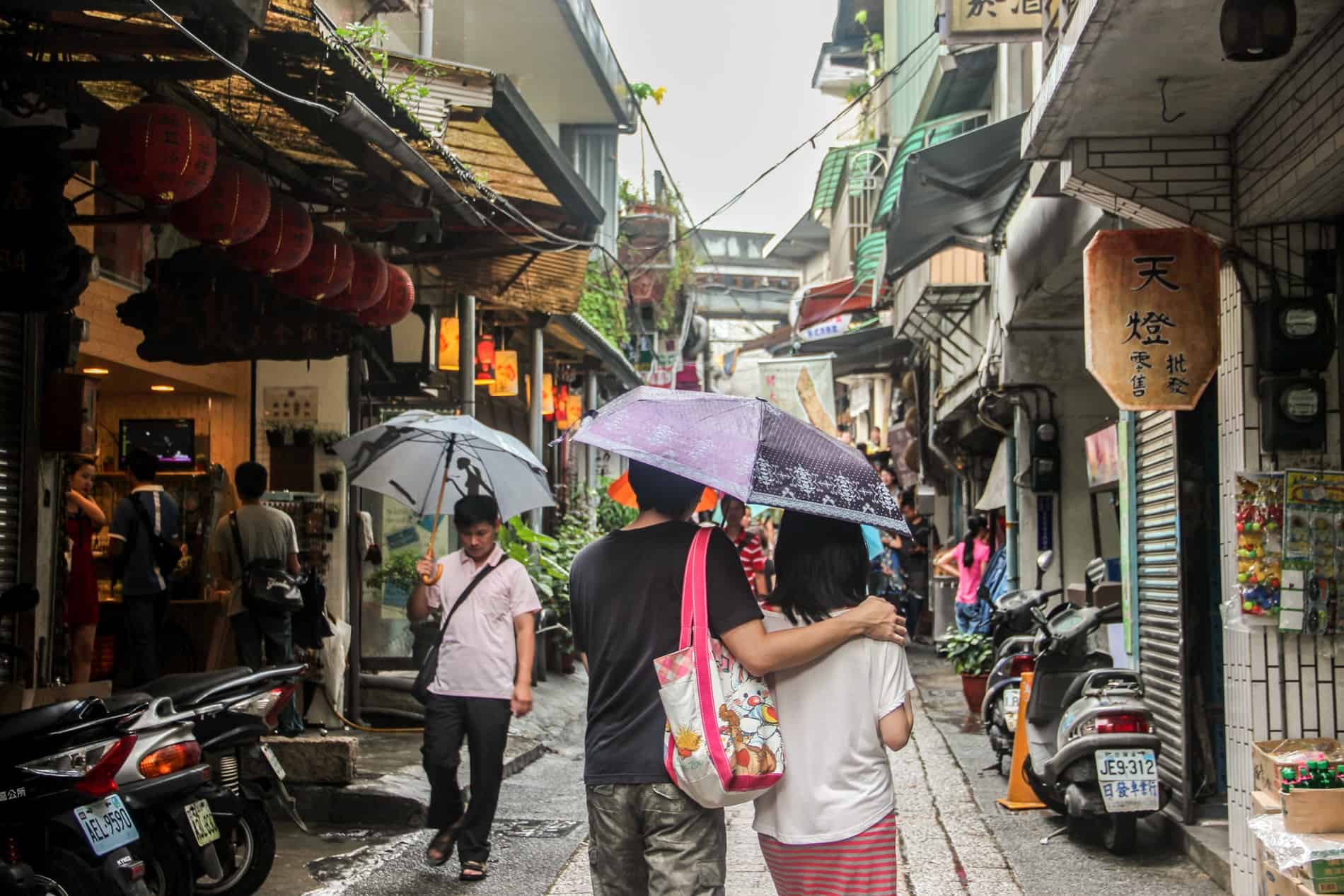 A couple walking under an umbrella down an old, narrow street of store-fronts and parked motorbikes. 