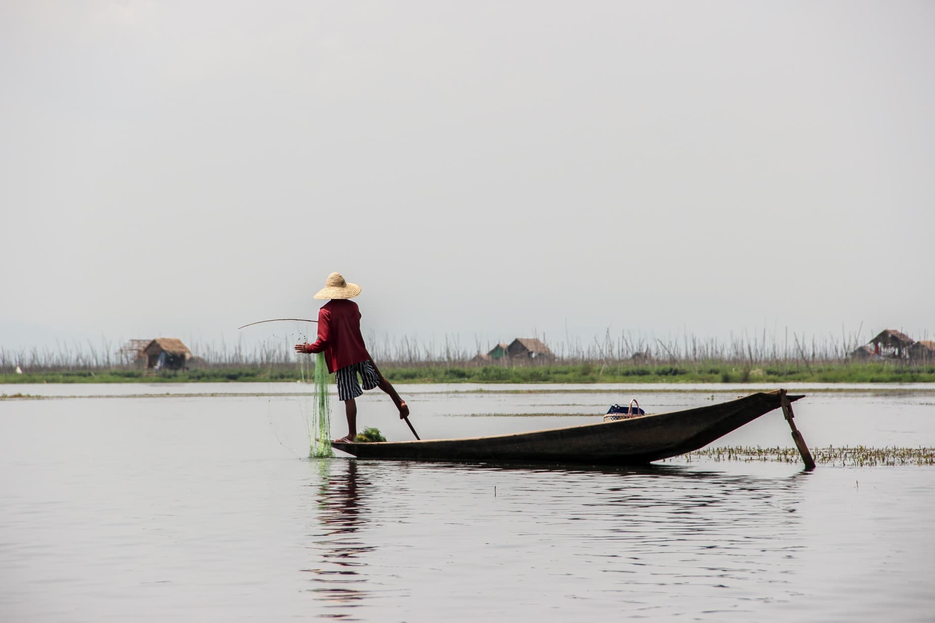 A fisherman on Inle lake standing up on his boat, dropping a green net into the water and rowing the oar with one leg.
