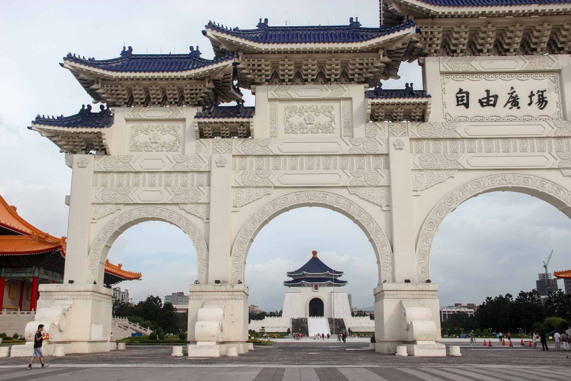 A white, motif carved archway with electric blue roof tiles, in front of a memorial hall in the same design. 
