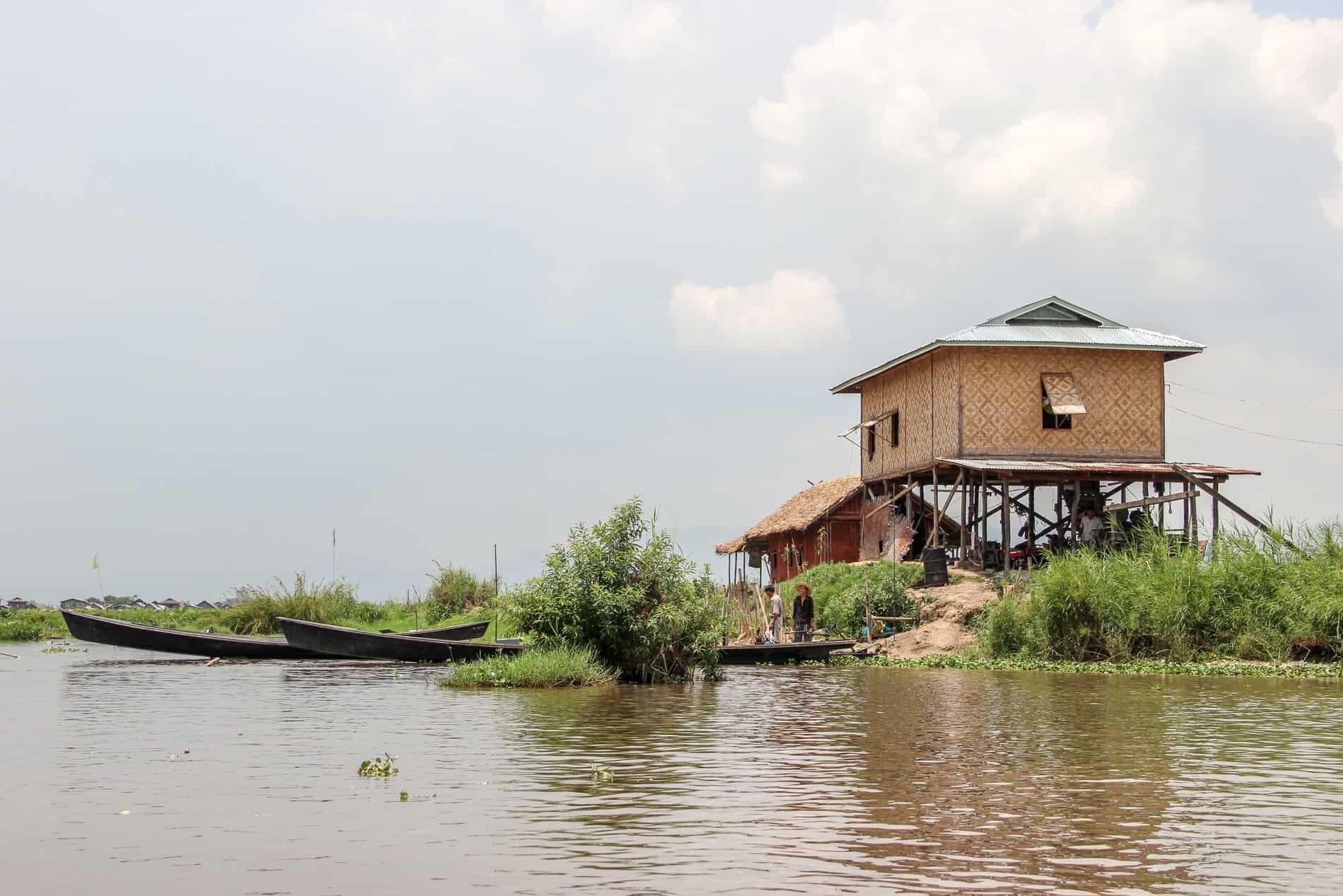 Two people standing at a dark longtail boat, in front of a large, caramel coloured bamboo stilt house. 