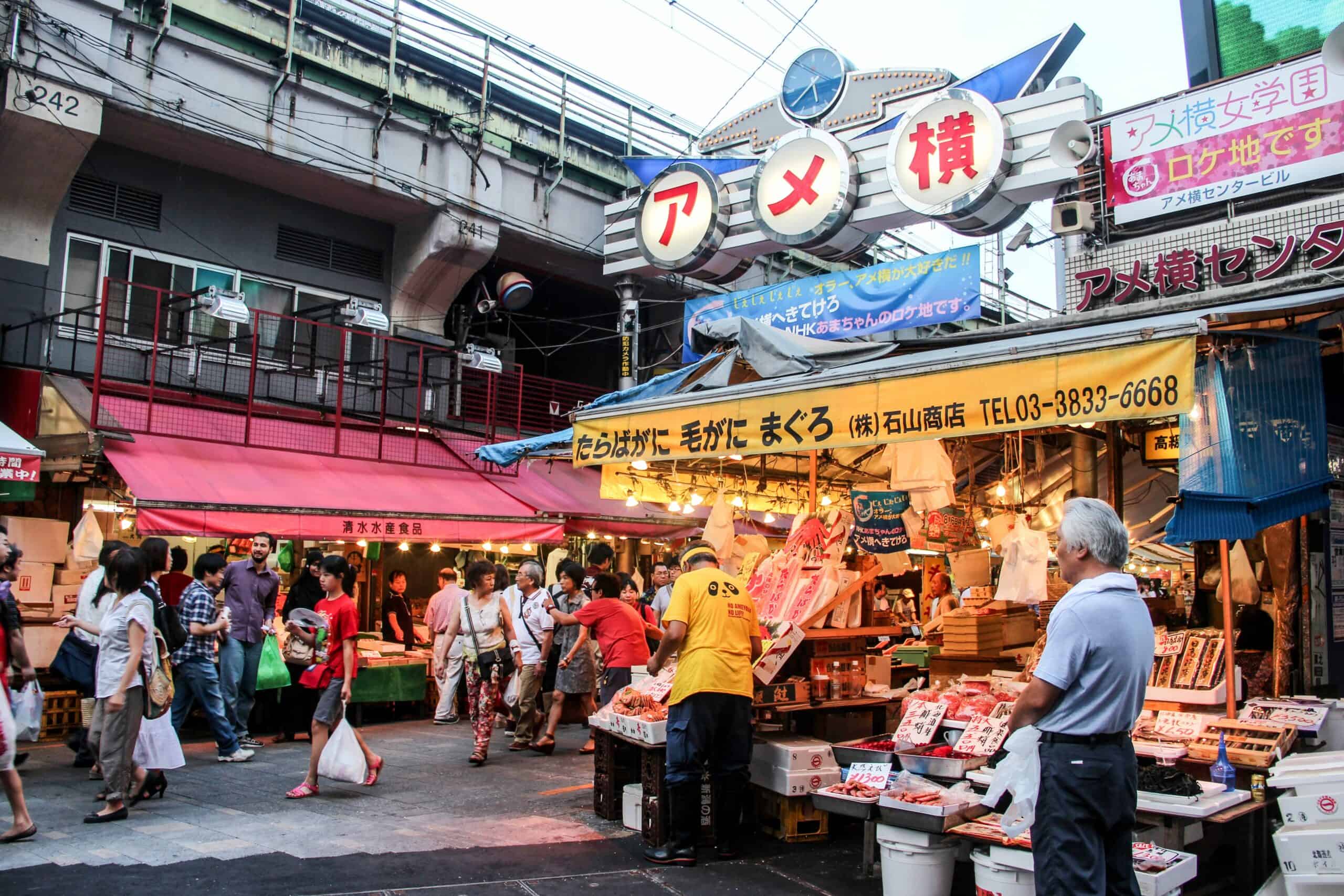 A busy street market with yellow and red shop fronts under a railway arch in Tokyo, Japan.