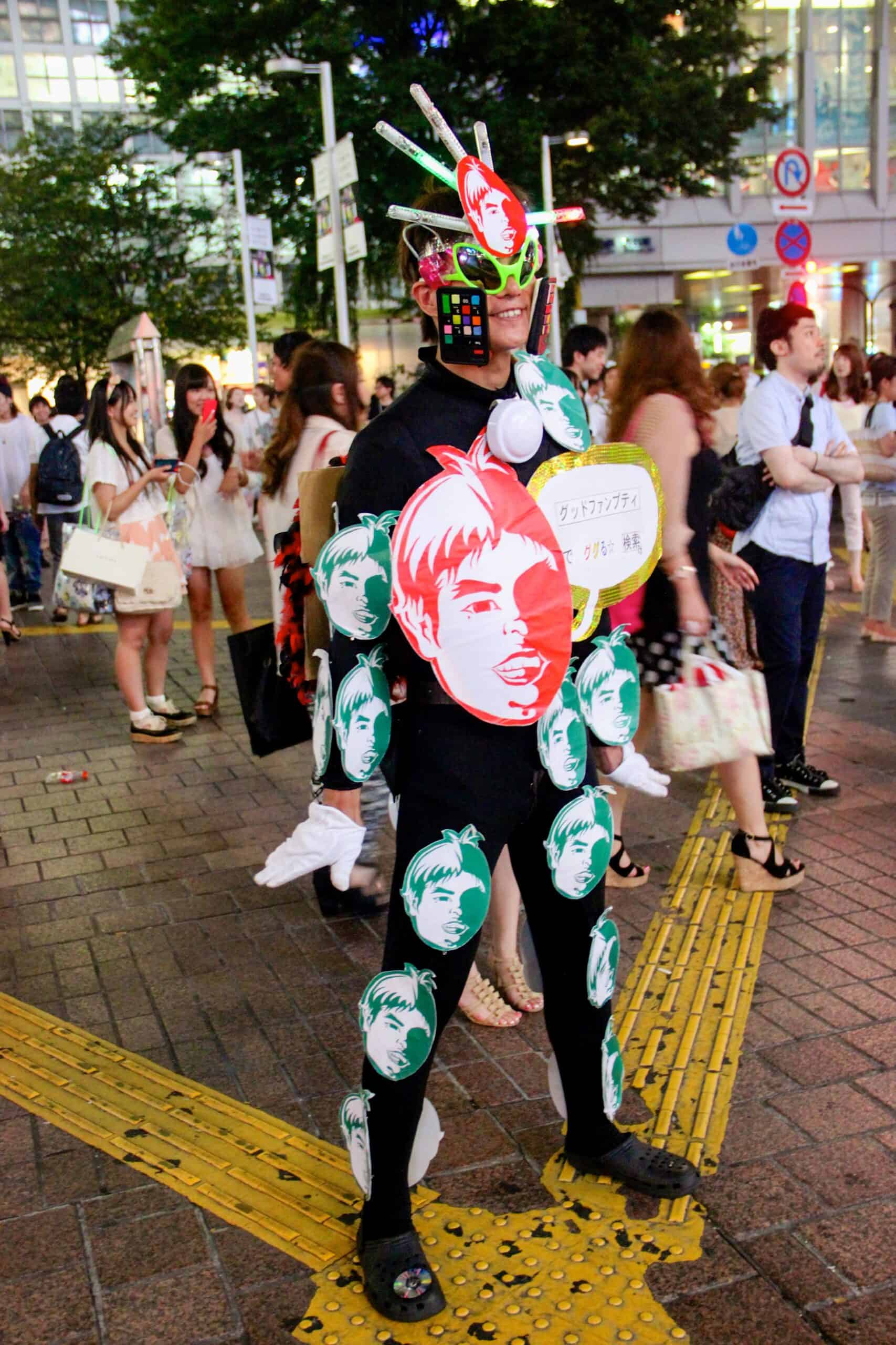 A Man in a costume of paper faces, lime green sunglasses and a pretend mobile phone stuck to his face on the streets of Tokyo. 
