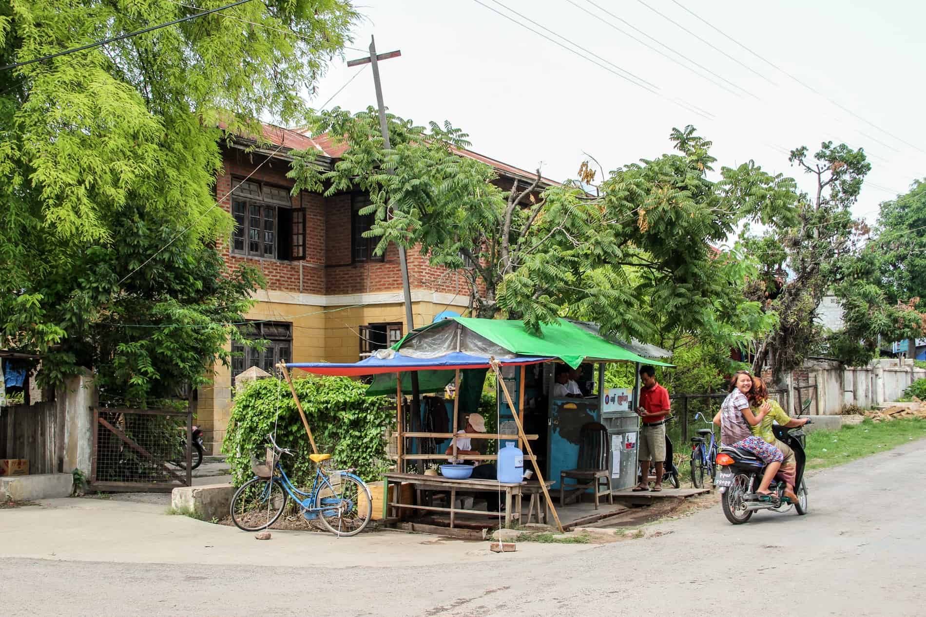 A woman on the back of motorbike smiles to the camera while passing a makeshift roadside stall with a bright green roof in the leafy Nyaungshwe Township. 