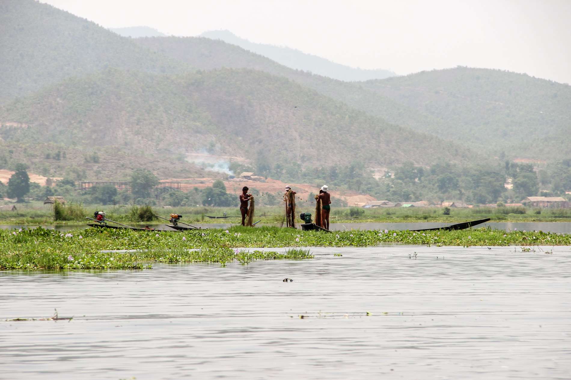 Three fishermen casting nets into the waters of Inle Lake next to a floating garden. Behind are the forest covered mountains. 