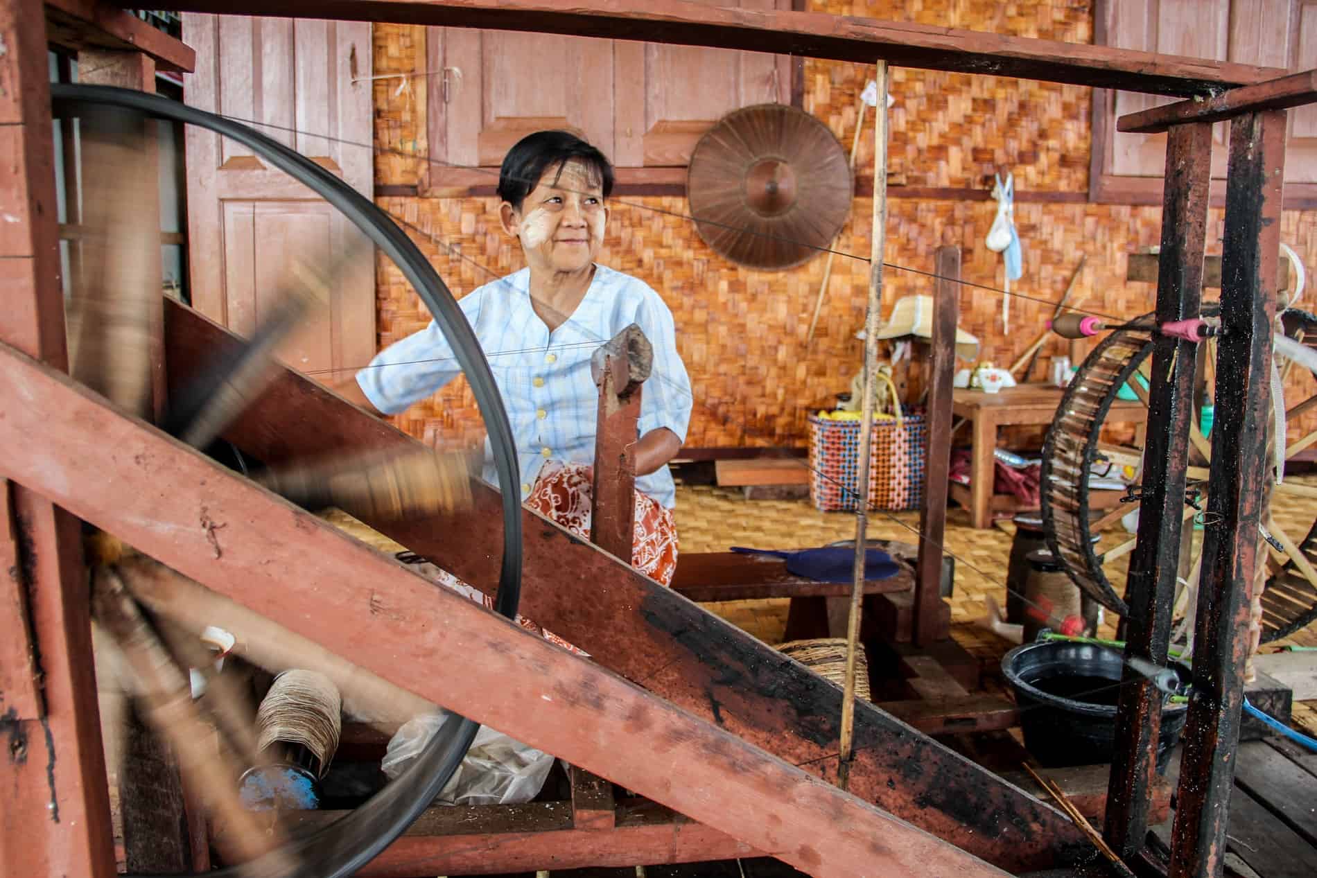 A Burmese woman with painted cheeks works a weaving machine in a bamboo house. 