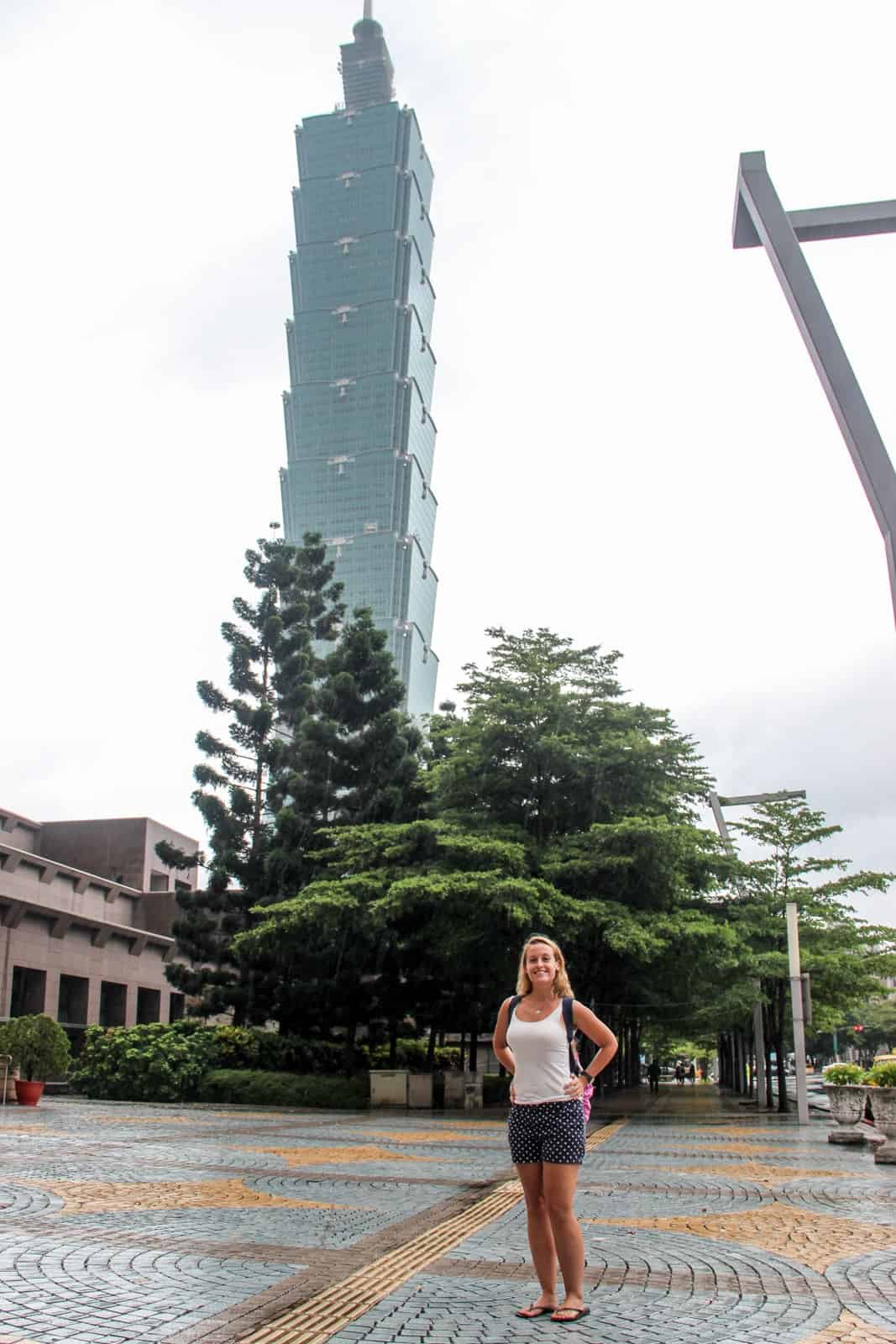 A woman stands in front of the Taipei 101 tower; a skyscrapper shaped like a bamboo plant. 