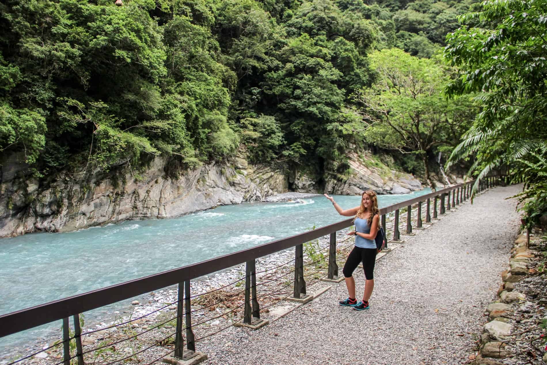 A woman points with both hands towards the bright crystal blue water of the Taroko gorge river and forested cliff. 