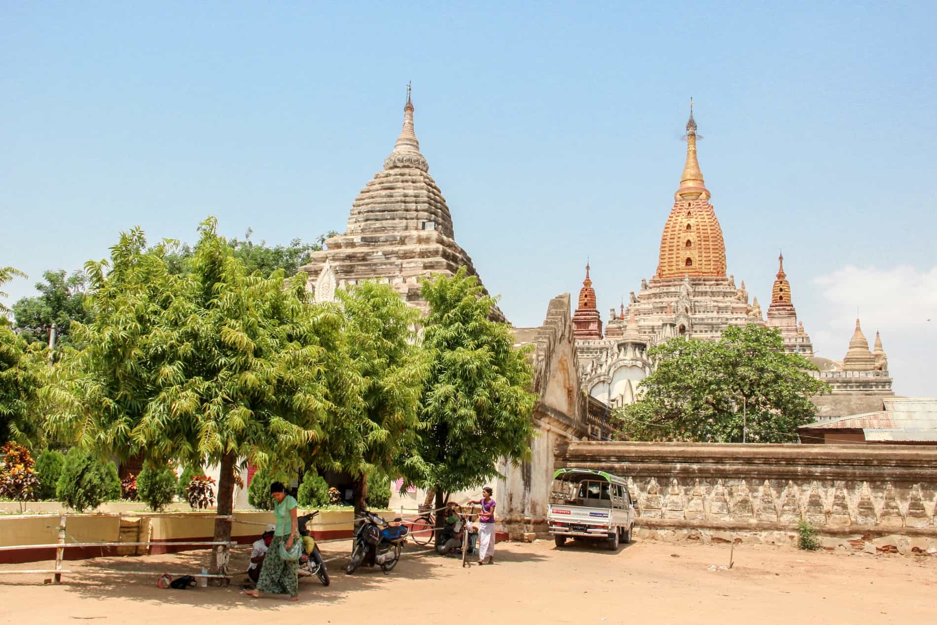 A woman pushes a bike in front of a tree at the entrance to a large temple complex with white and gold pagodas, in the dusty orange grounds of Bagan, Myanmar.