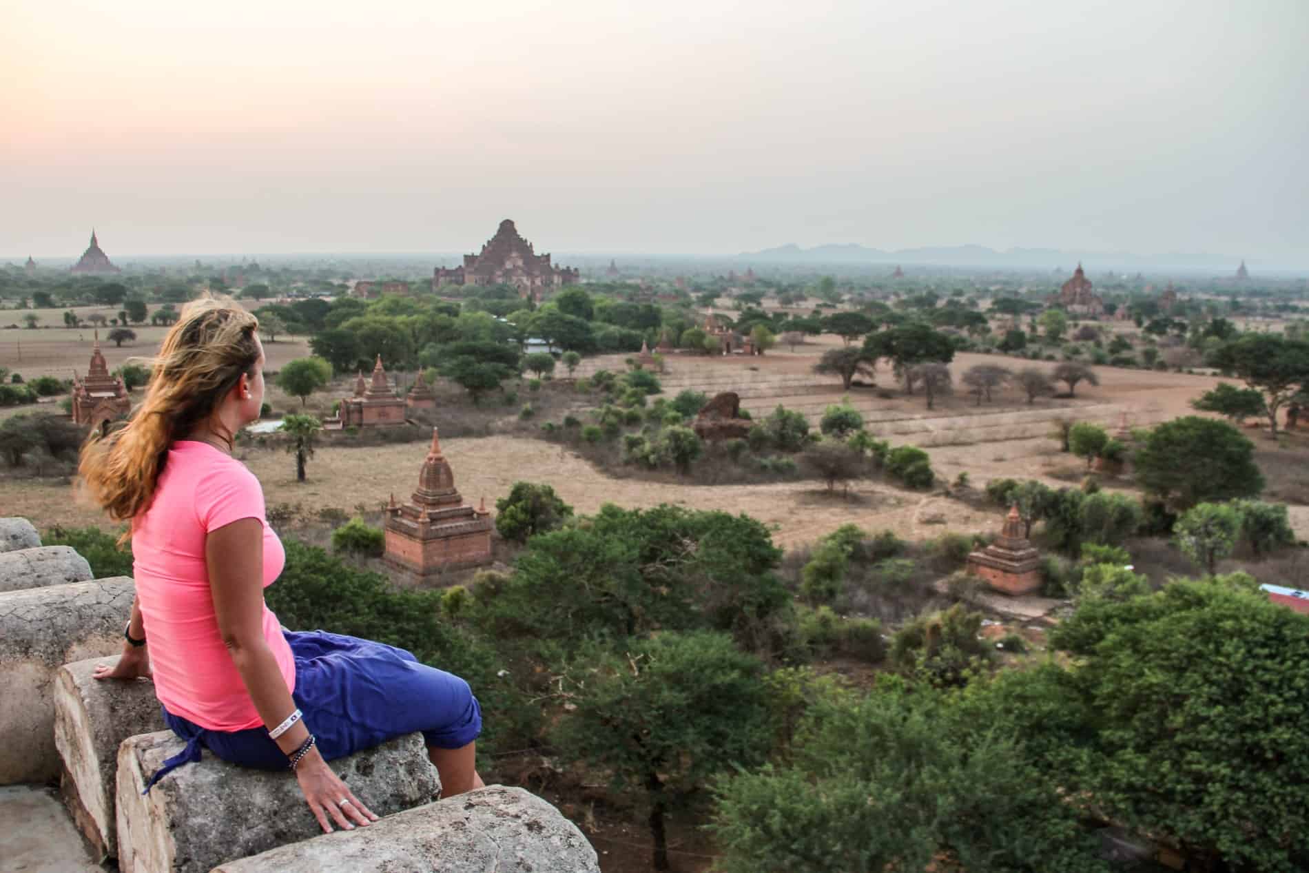 A woman in a neon pink t-shirt and purple pants sits on an arched stone watching sunrise from the top of a temple in Bagan, Myanmar looking out to a field scattered with golden brown temples poking through the grass. 