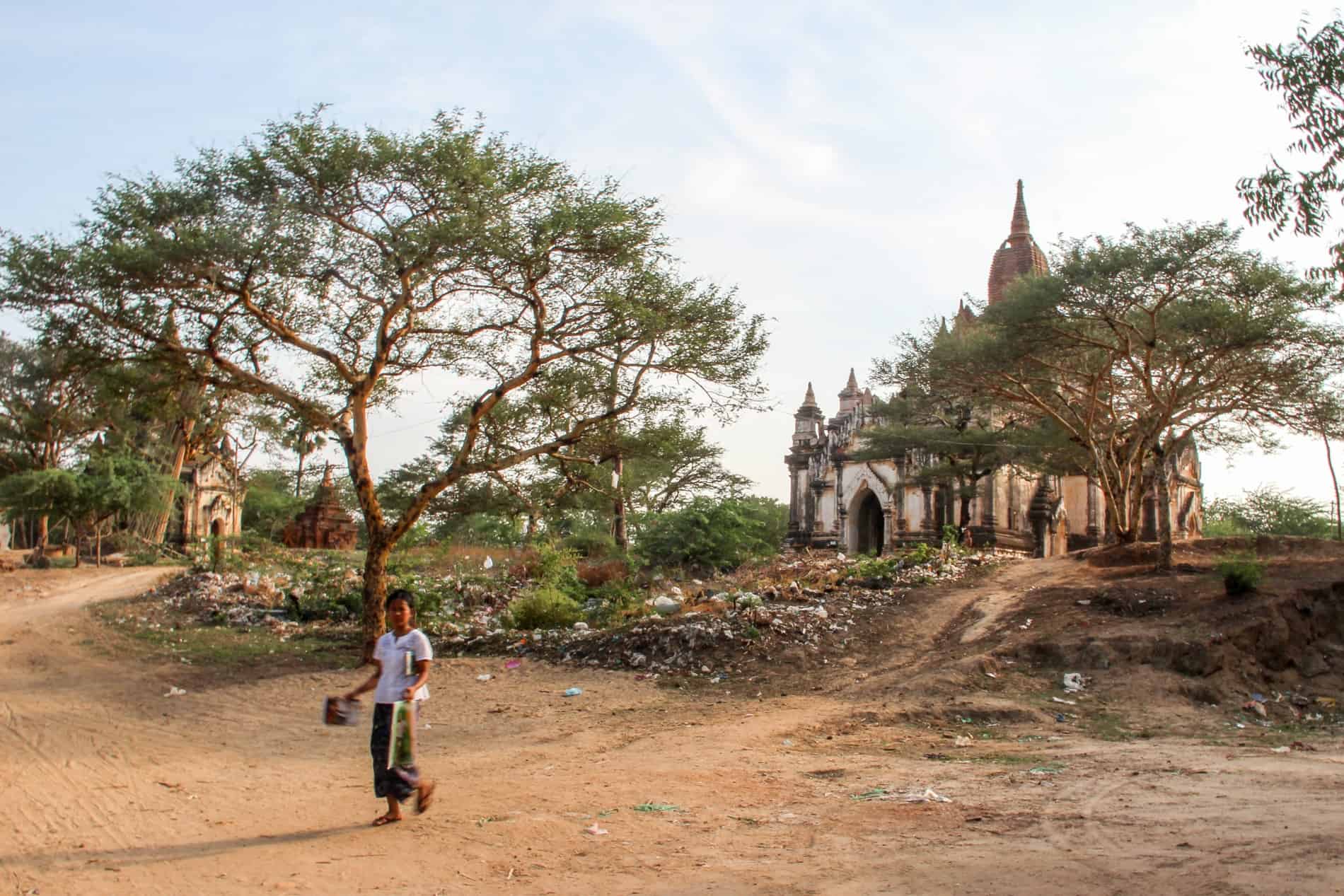 A Burmese woman on the dusty, arid roads of old Bagan outside a small off-white temple complex. 