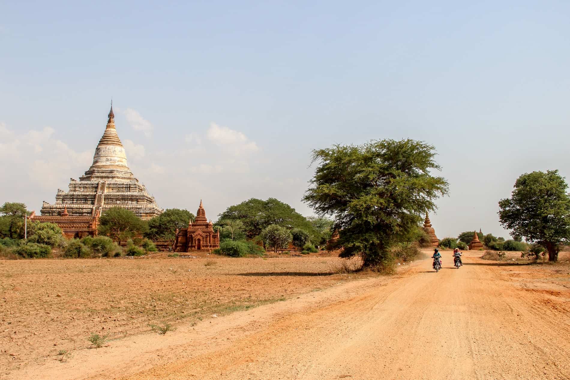 Two motorbikes riding on a yellow dusty road past a white temple pagoda. 