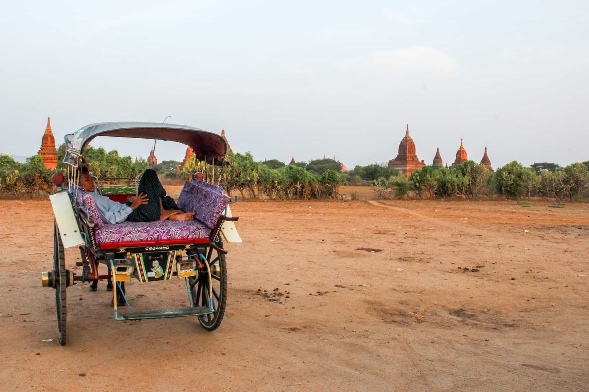 A male horse and cart driver rests in the carriage of his horse cart in front of a scattering of temples on orange dusty ground in Bagan, Myanmar.