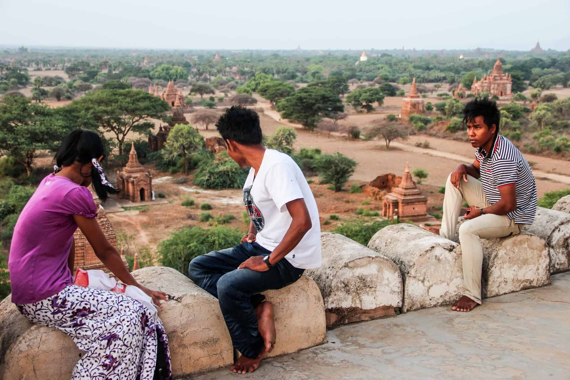 Three Burmese locals sitting on a stone temple wall overlooking the vast tree and temple-dotted plain. 