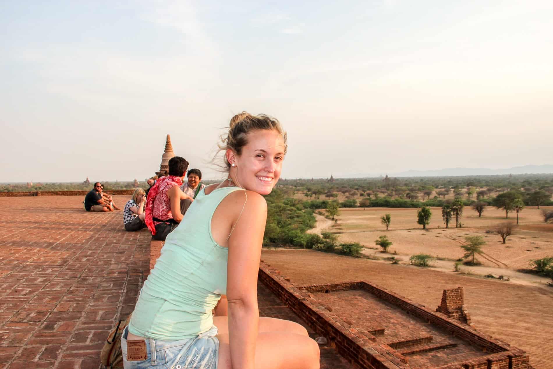A woman looks towards the camera whilst sitting on an elevaed brick temple wall overlooking the vast temple grounds of Bagan in Myanmar. 