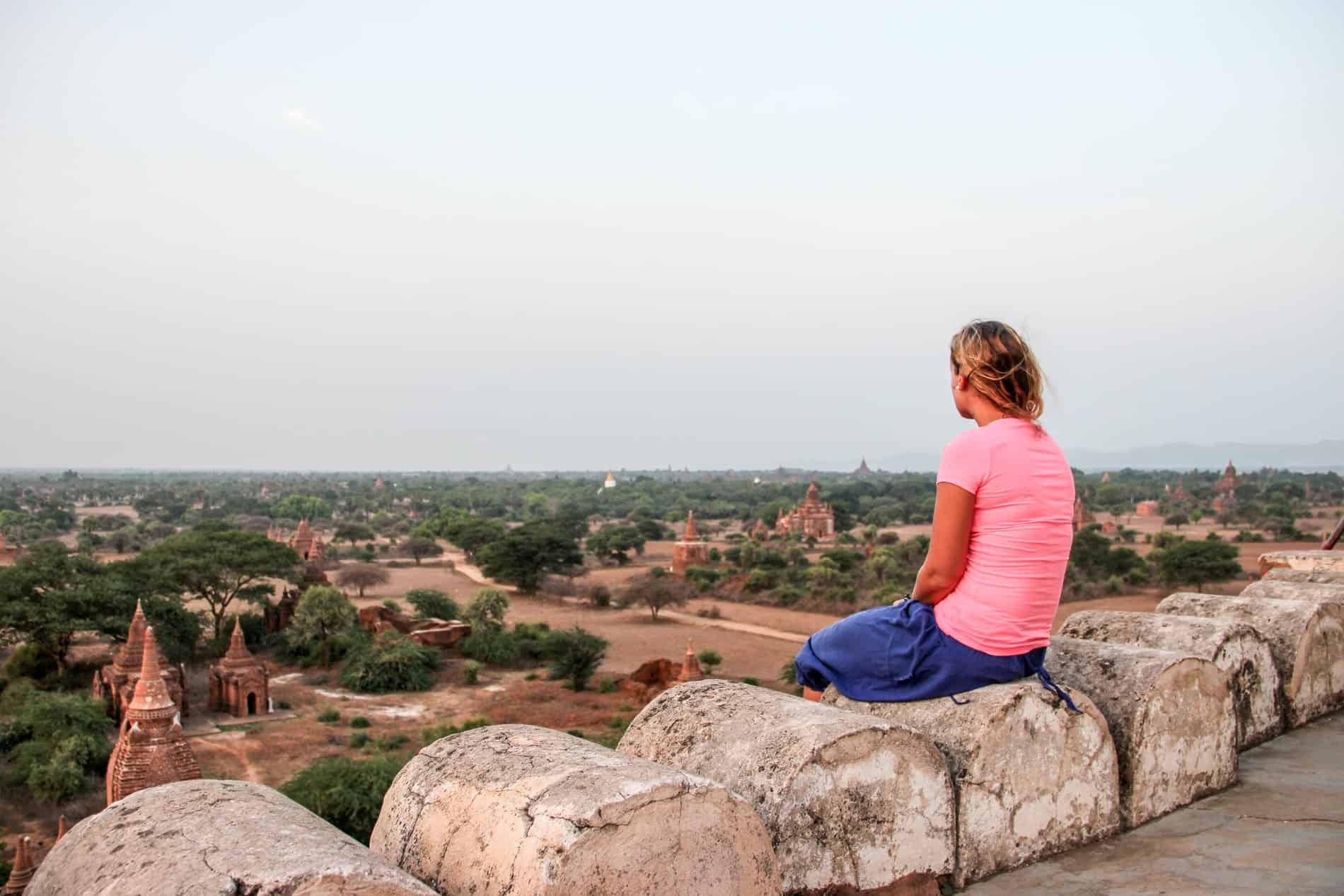 A woman in a neon pink t-shirt and purple pants sits on an arched stone watching sunset from the top of a temple in Bagan, Myanmar. In the fields ahead are a scattering of golden brown temples poking through the grass. 