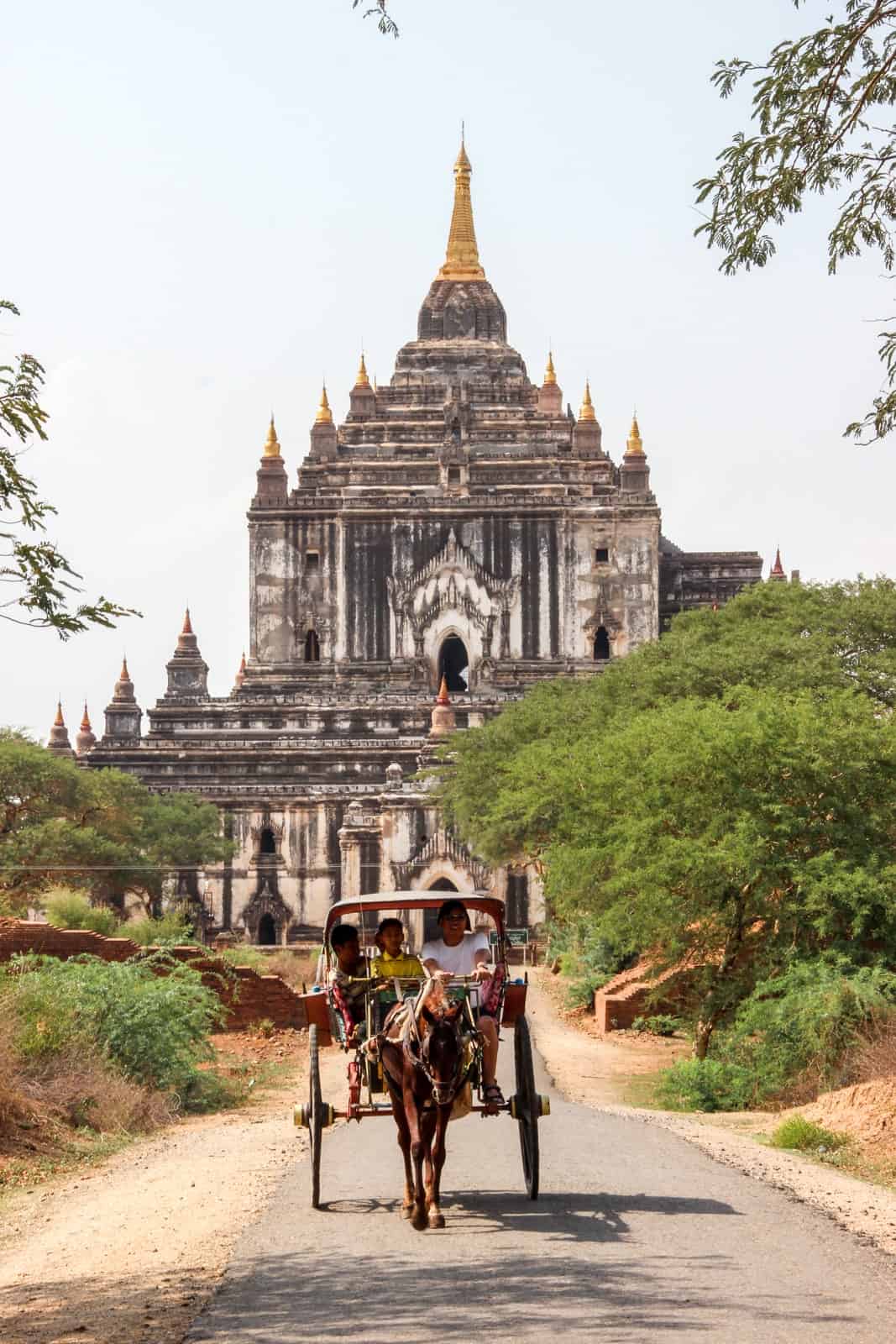 A woman and two children riding in a horse drawn cart in front of a tiered, gold spired temple in Bagan. 