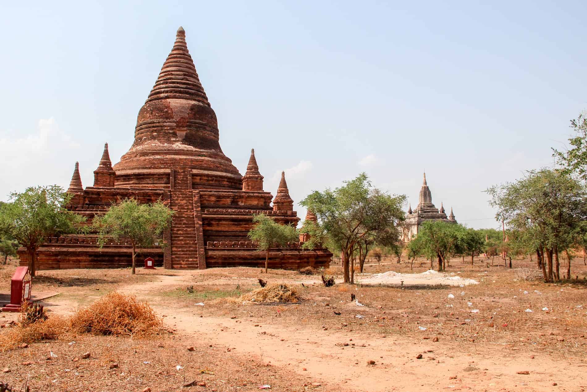A large ochre red brick pagoda in front of a white temple in an isolated dusty forest. 