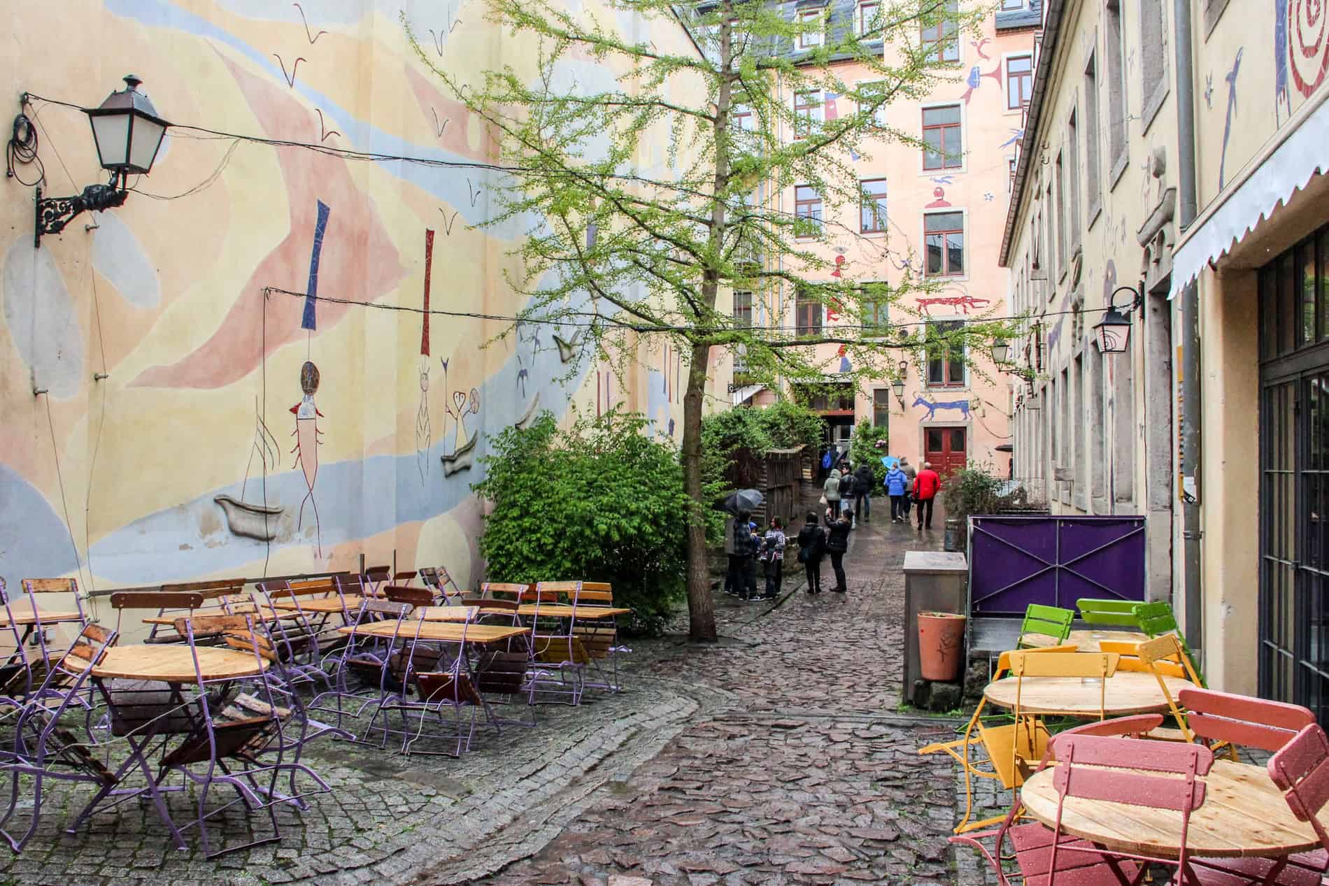 People walking down a cobblestone street with colourful tables and chairs and walls painted with motifs of animals. 