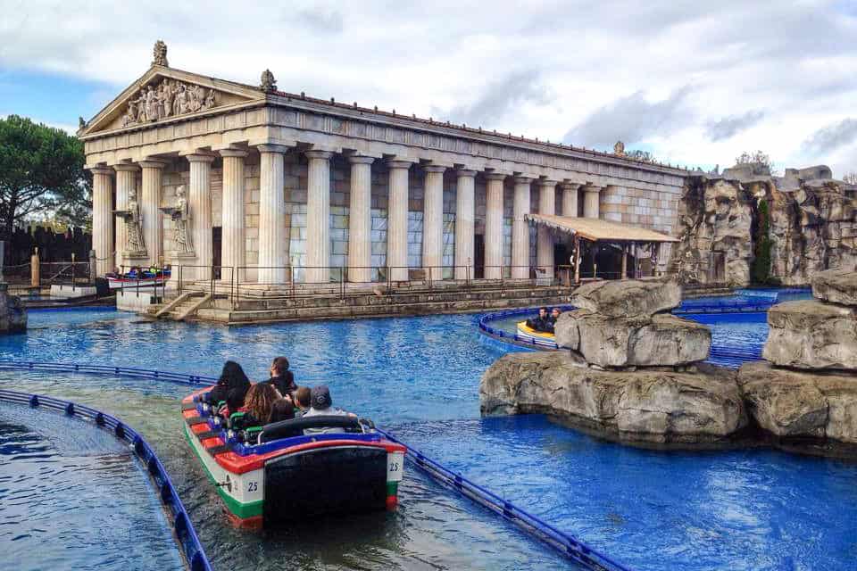 A theme park water boat ride floats past a replica Greek marble, columned building at a theme park in Germany. 