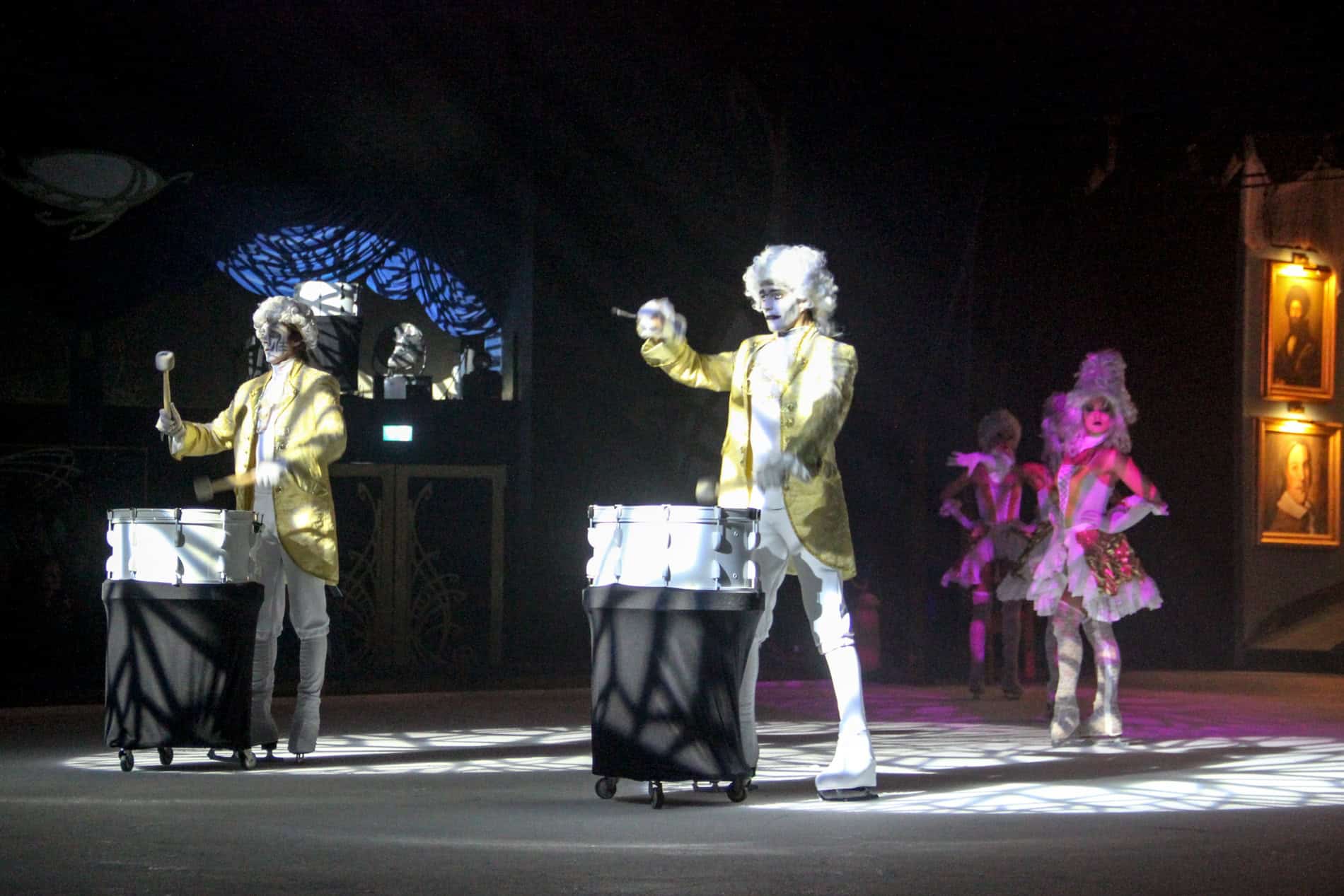 Two ice skaters with white wigs and yellow jackets playing the drums. 