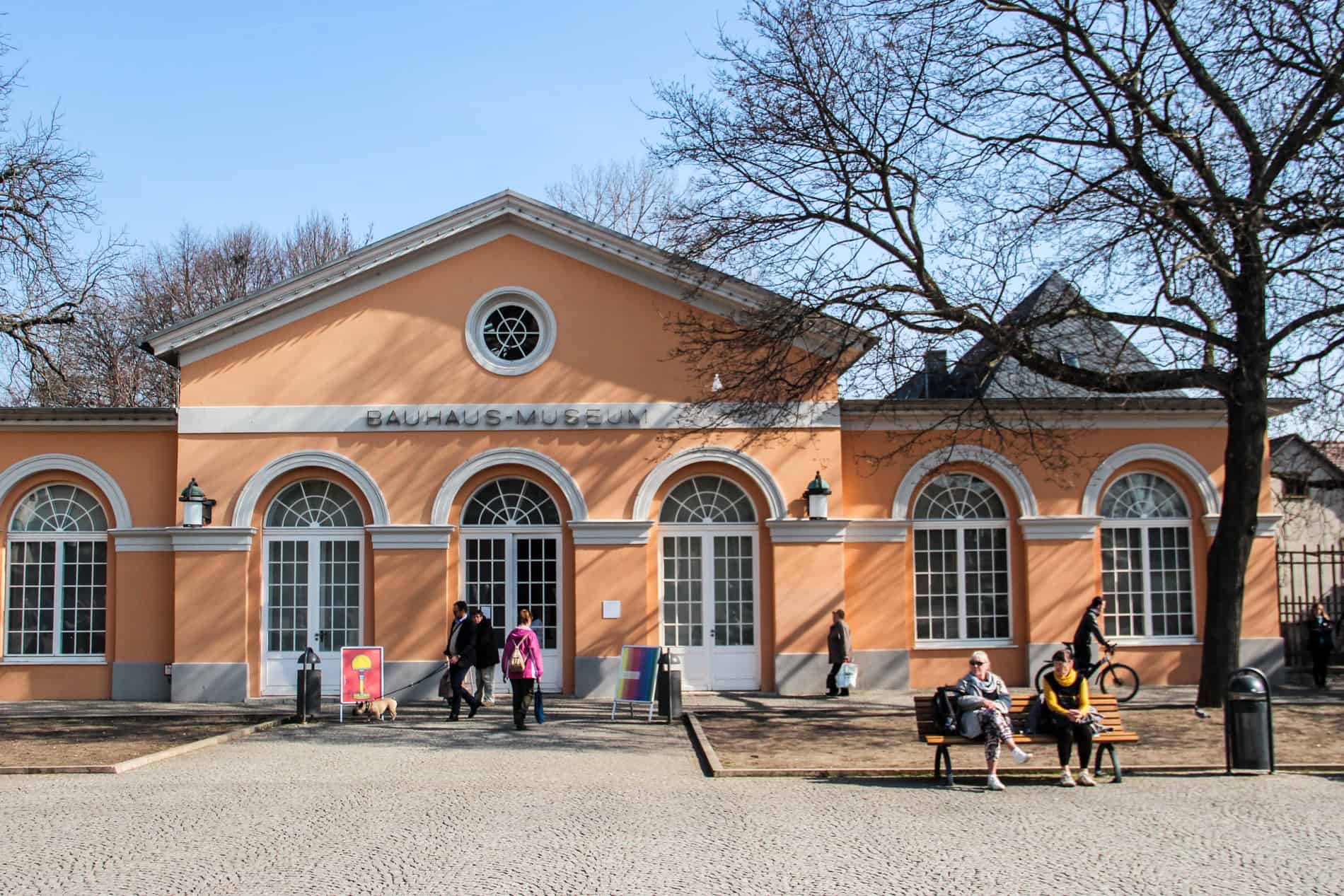 People sitting on a bench outside of the orange Weimar Bauhaus Museum.
