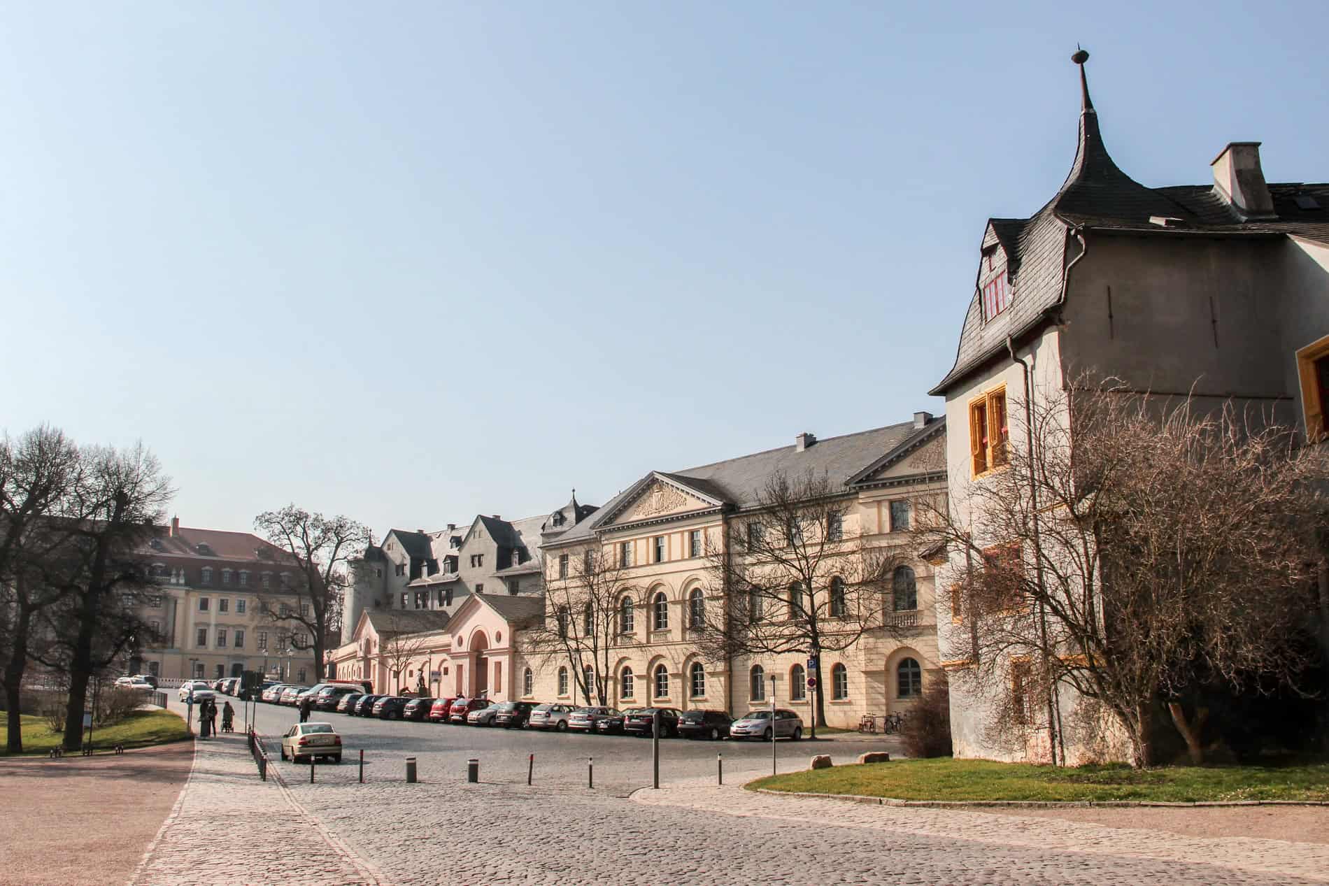 A wide cobblestoned street running past a long, beige baroque castle complex in Weimar, Germany. 
