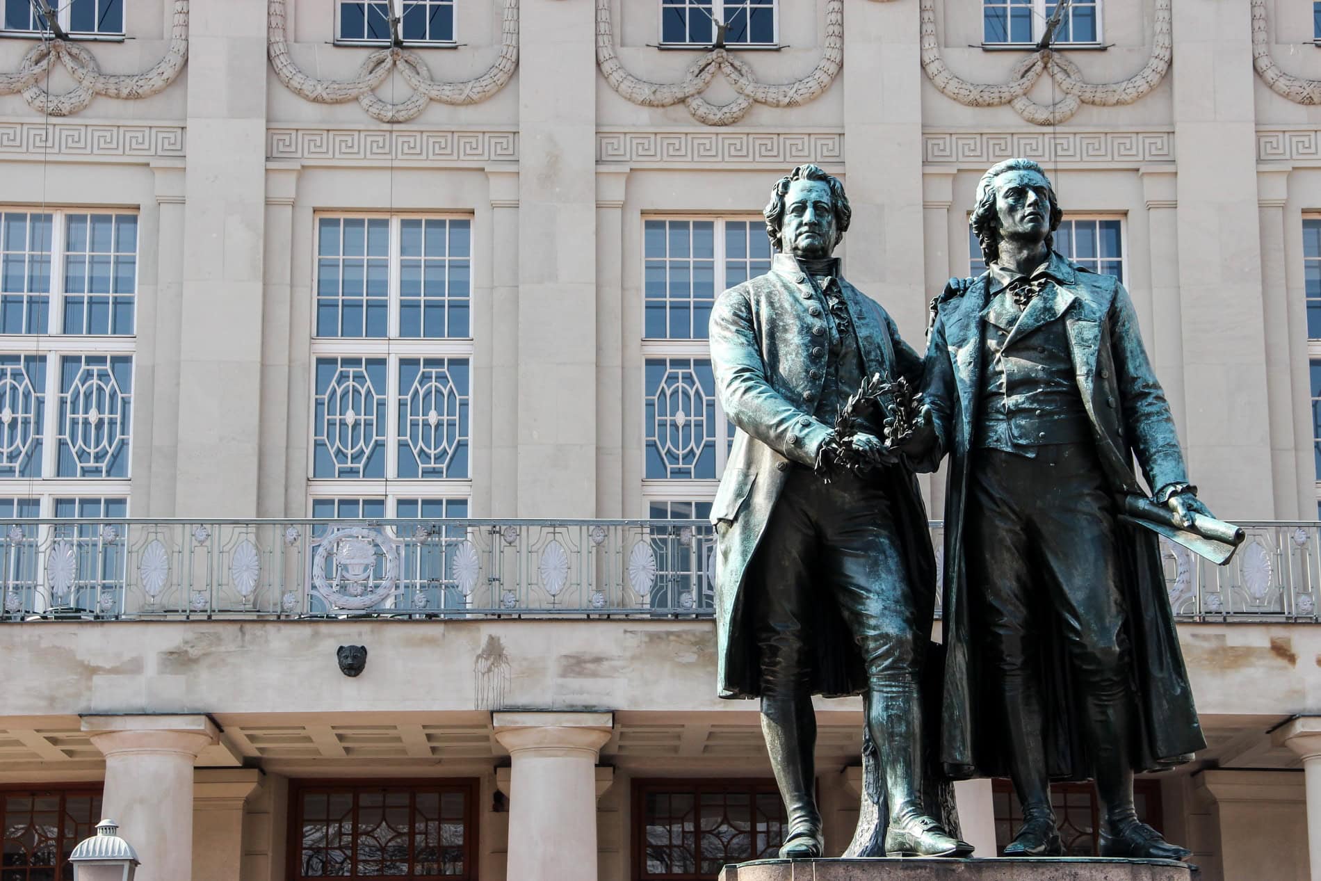 Close up of two blue-copper statues of Goethe and Schiller in front of a grand building in Weimar. 