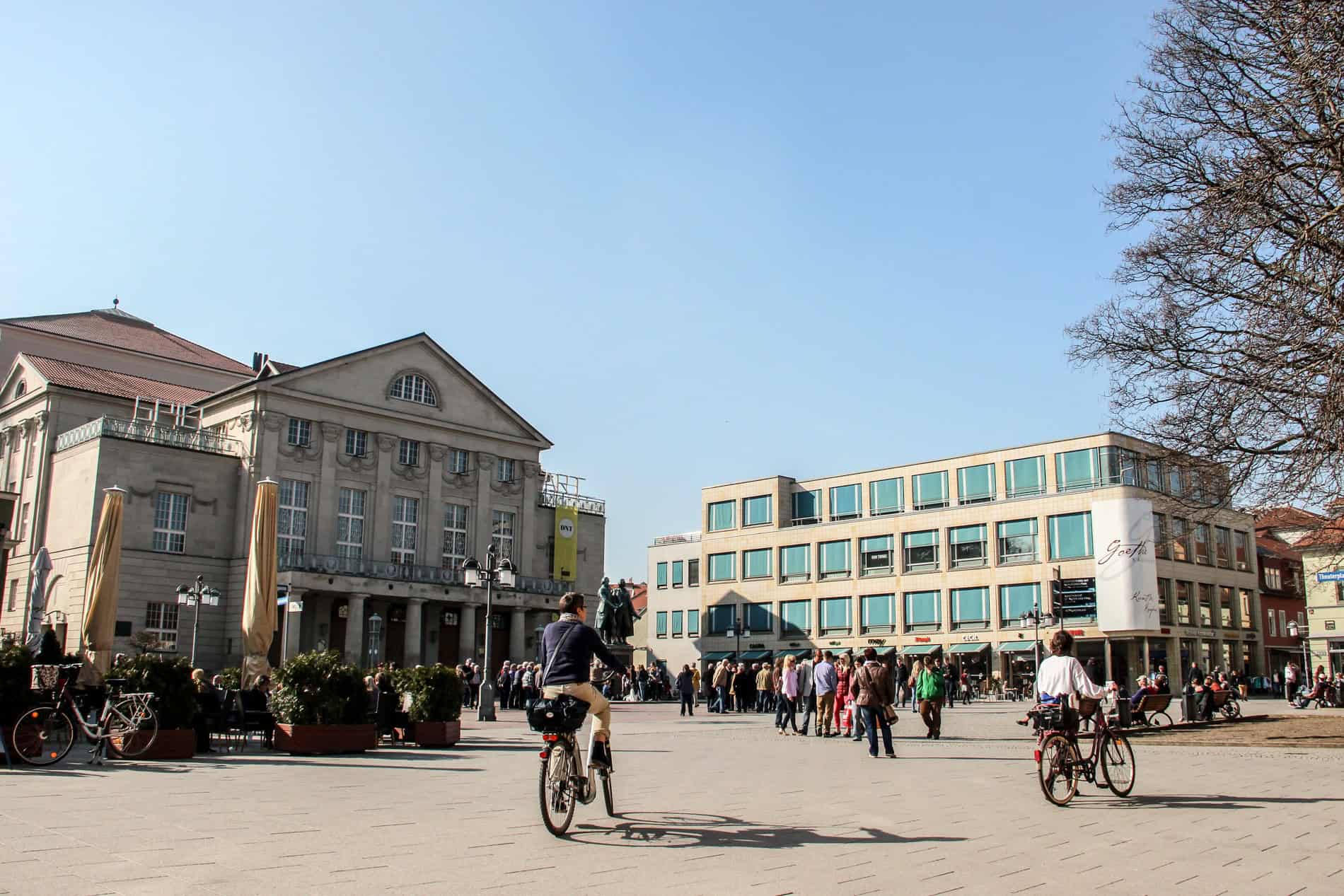 People riding bikes through a large public square in from of an old theatre and a modern blue-block glass building. 