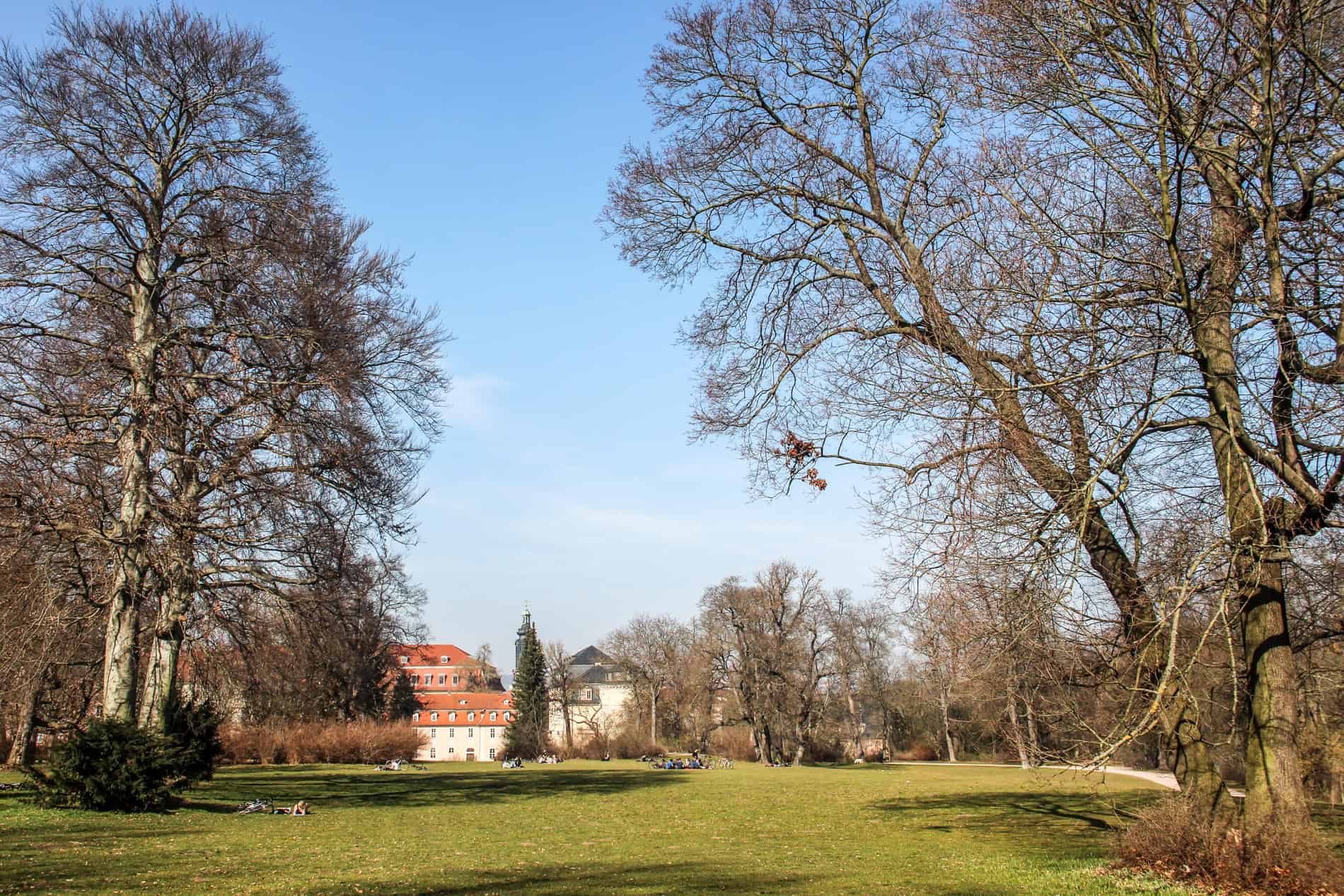 People lounging on the lawn of a huge park in front of a orange-roofed manor house. 