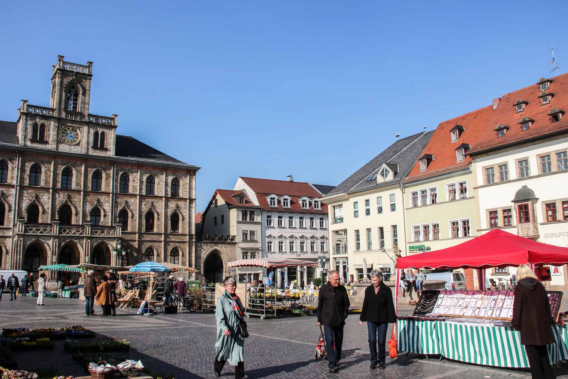 People walk past colourful stalls in a market square surrounded by gothic and baroque buildings. 