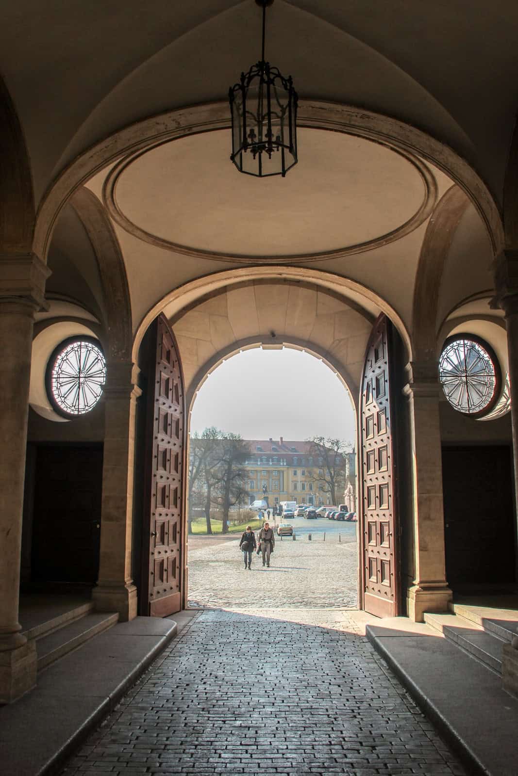 Two people walking towards a grand doorway in an arcaded roofed room. 