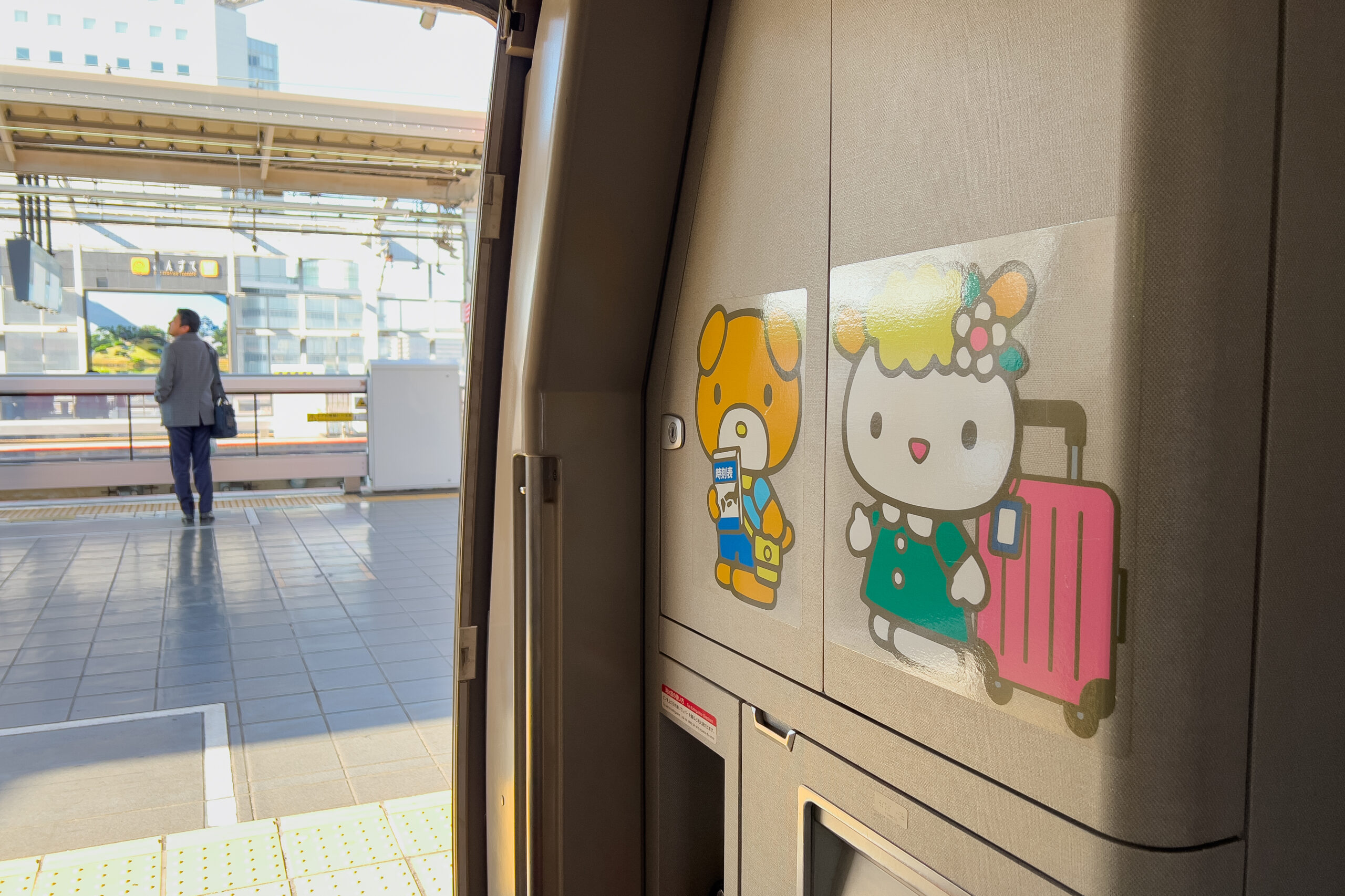 From from inside a train decorated with two Sanrio character stickers, looking towards the platform where a man is standing.