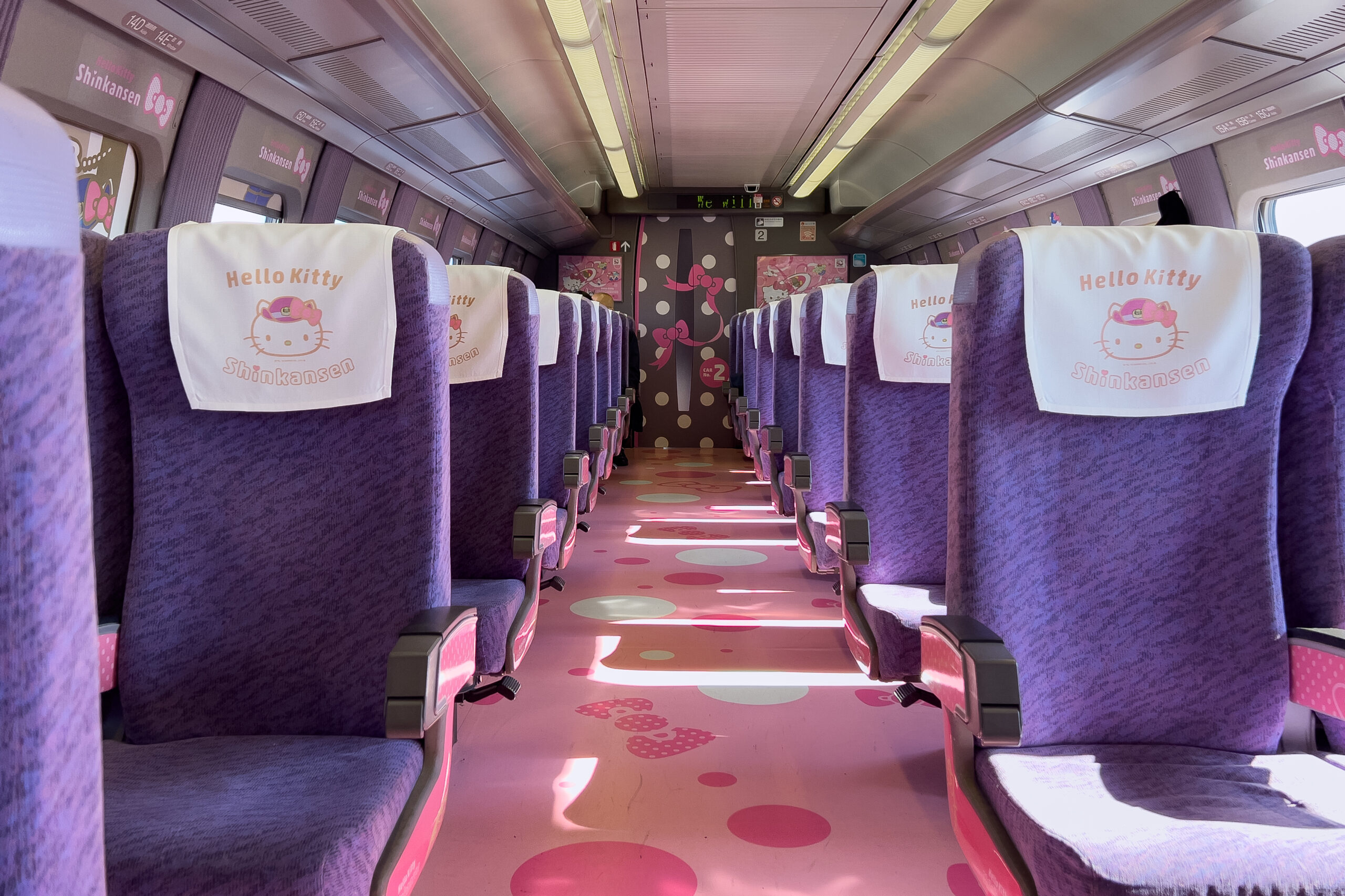 Inside view of the playful Hello Kitty Shinkansen Kawaii! Room carriage interior featuring bright purple seating with Hello Kitty headrest covers, a bow motif pink floor, polka dot door and window graphics.