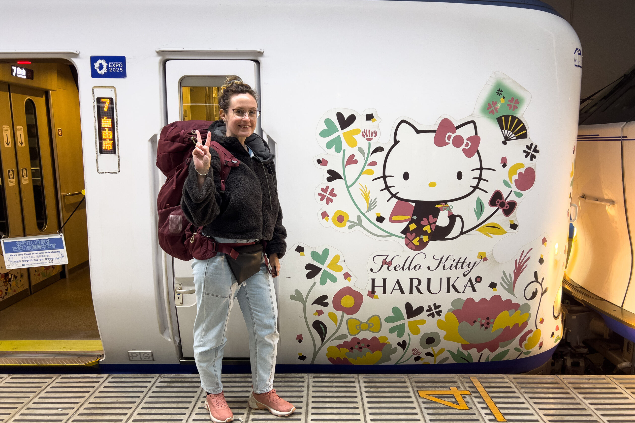 A woman on a train platform giving the peace sign stands outside the themed The Hello Kitty Haruka Kansai Airport Express train.