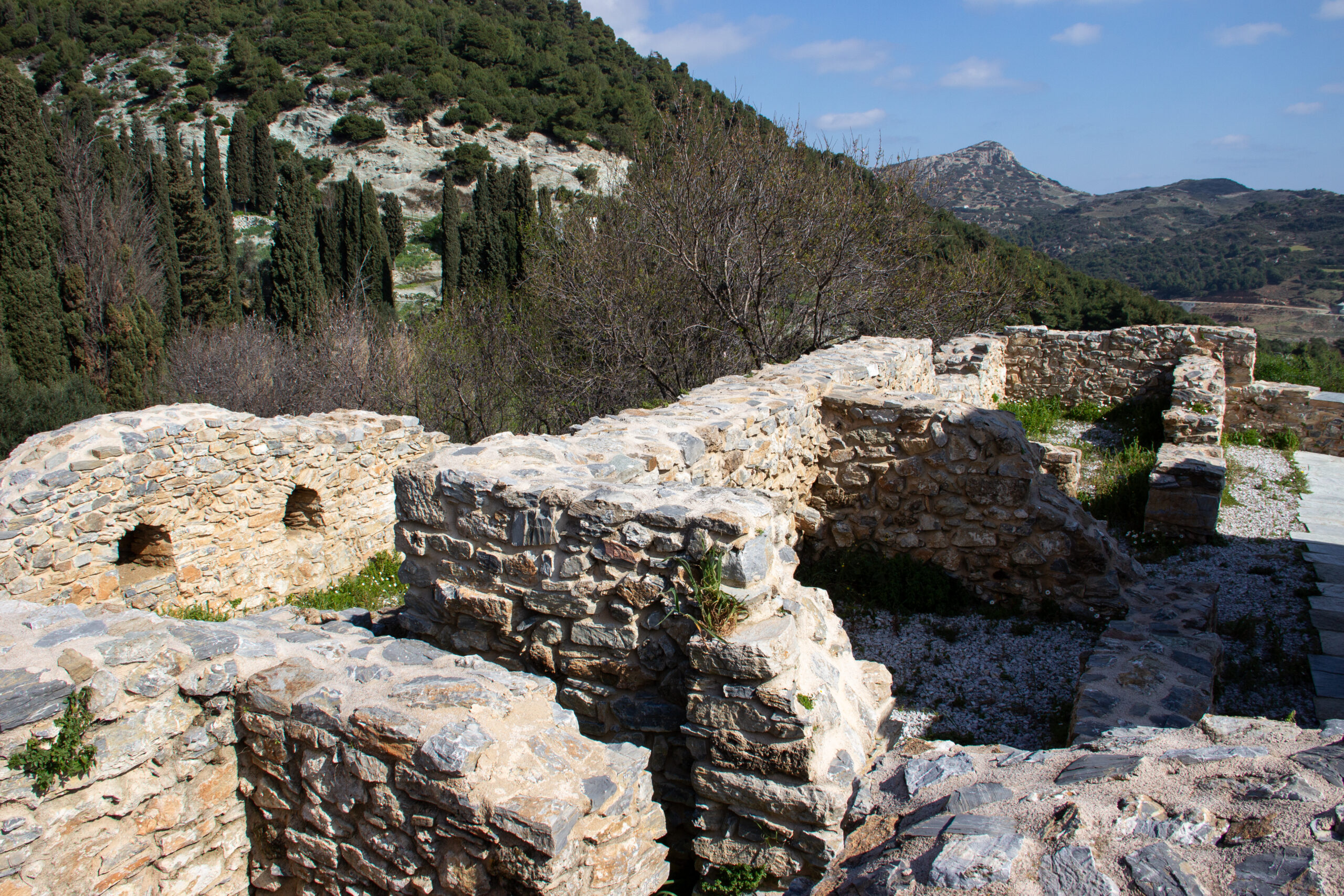 A cluster of stone ruins on a green ledge overlooking forested hills and low mountains.