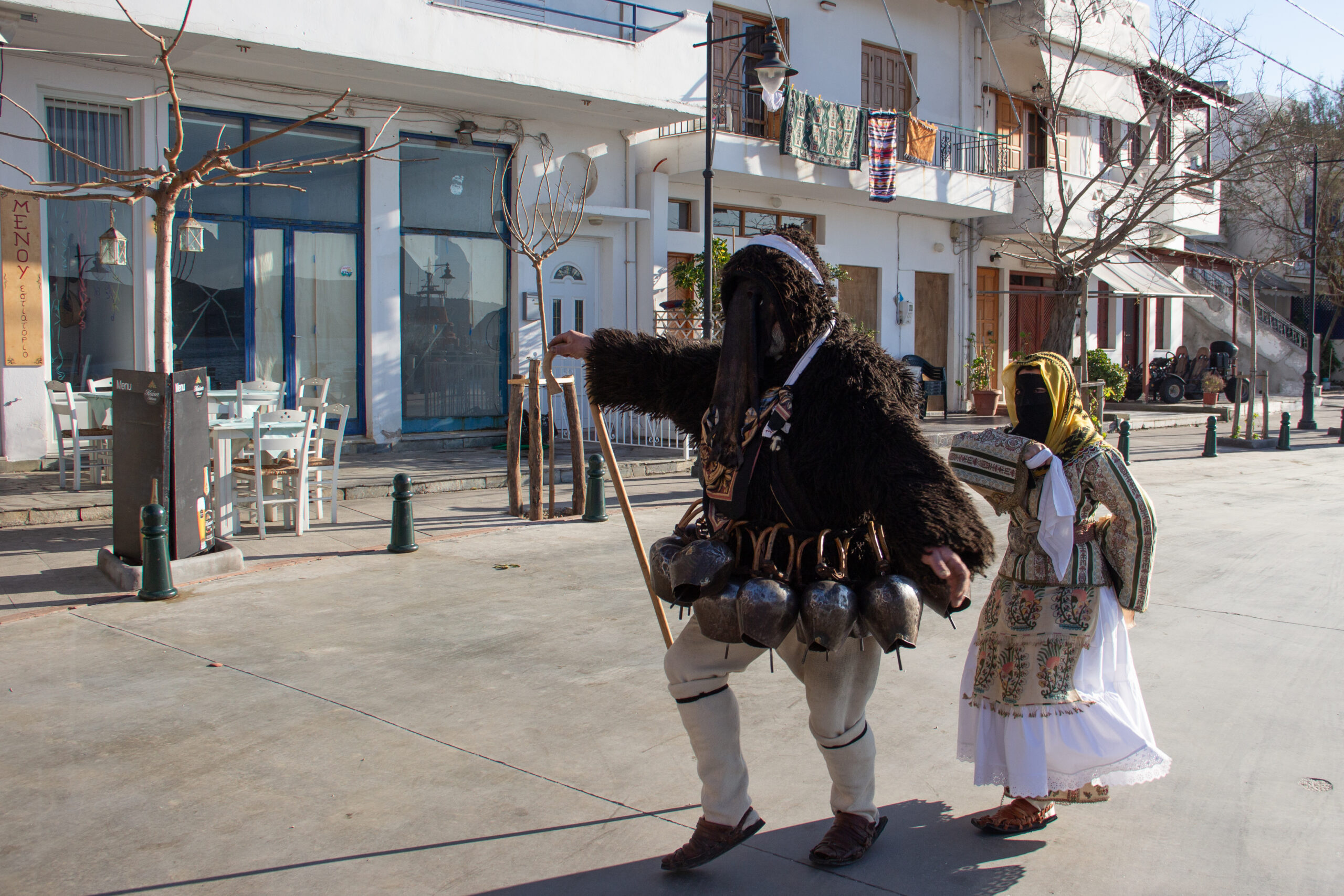 Two costumed figures in Skyros Carnival outfits – the goatman in a shaggy black coat with goat bells and a veiled woman waving a white handkerchief – performing in the street.
