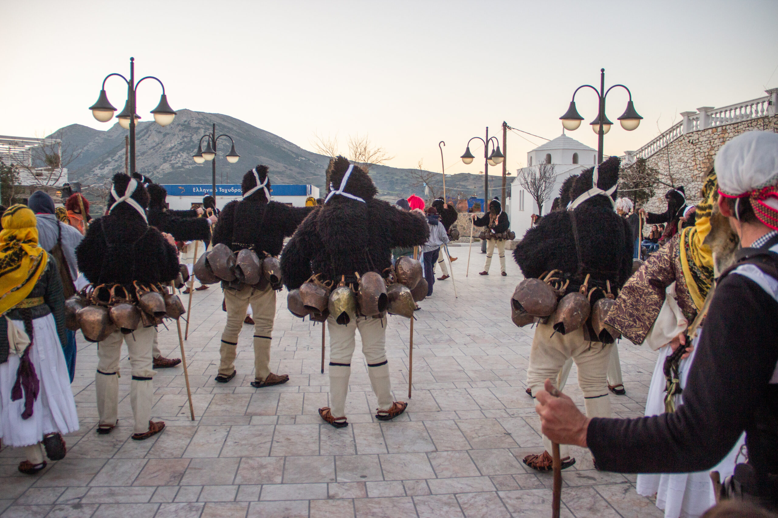Skyros carnival participants gathered on a street to a backdrop of a white church and distant hills.