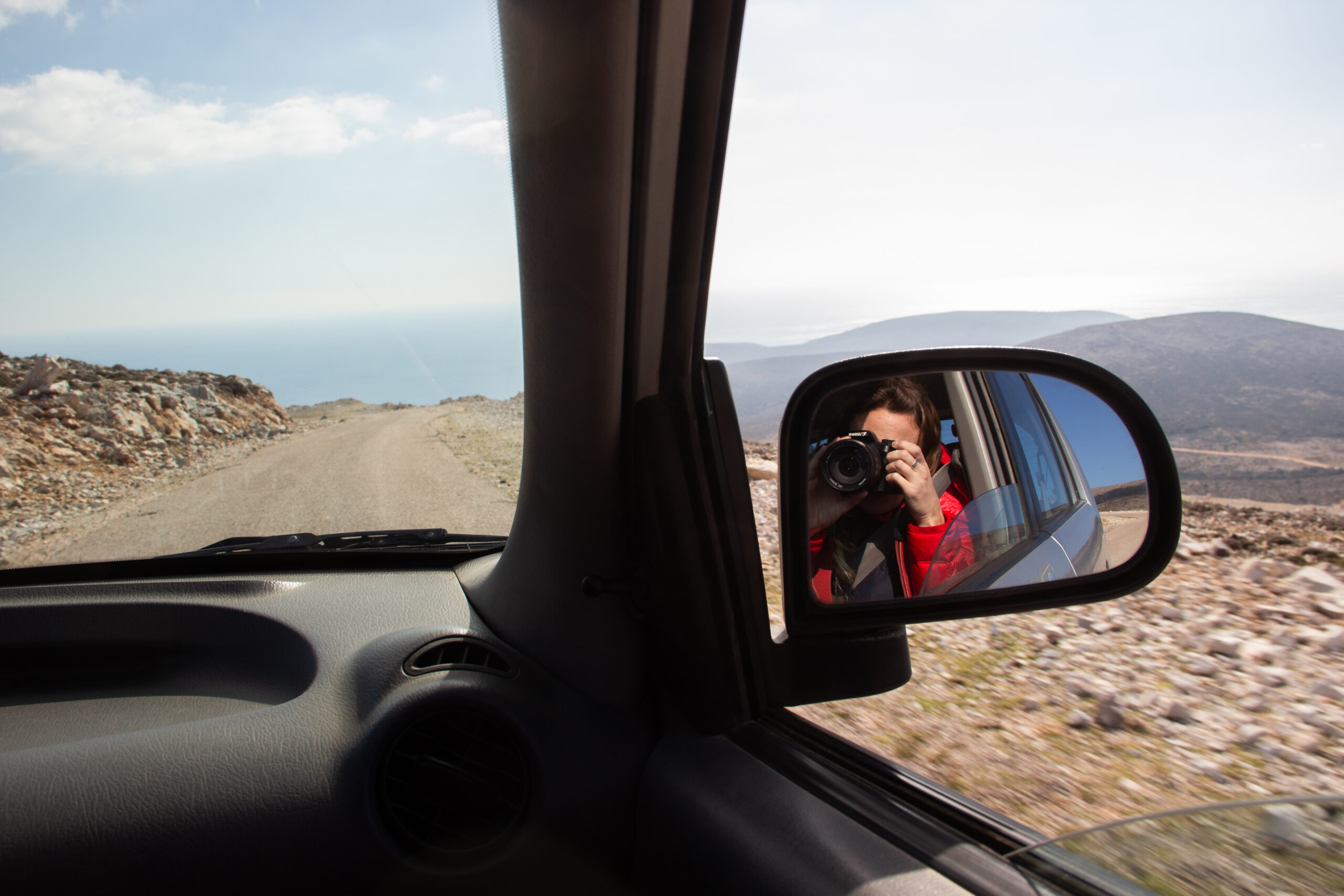 Reflection of a woman in a red coat and holding a camera in the car's side mirror while driving through a scenic dry and rocky landscape.