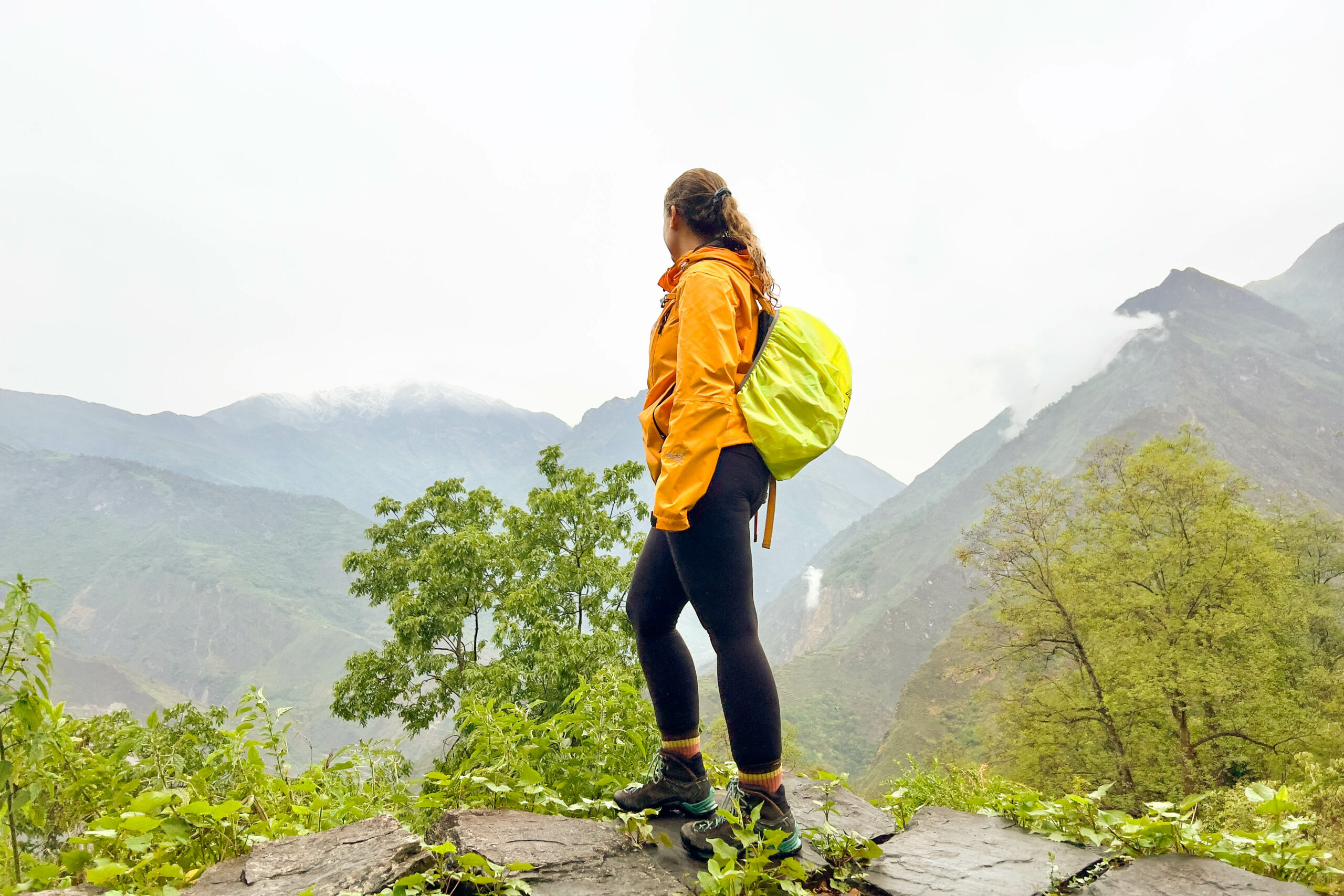 A female hiker in an orange jacket and lime green backpack stands on a stone ledge, looking out over foggy, green mountain peaks.