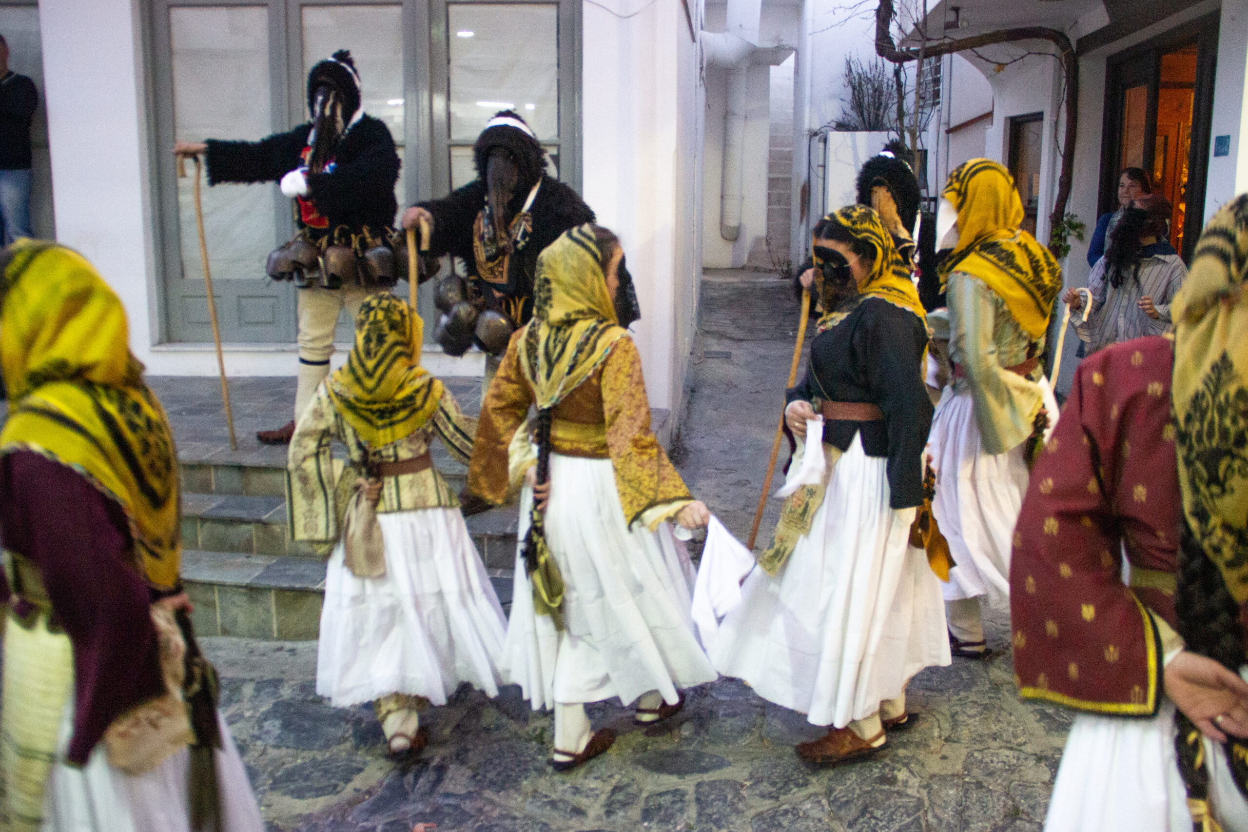 Masked carnival participants in Skyros performing in narrow town streets at dusk with wooden sticks and white cloth.