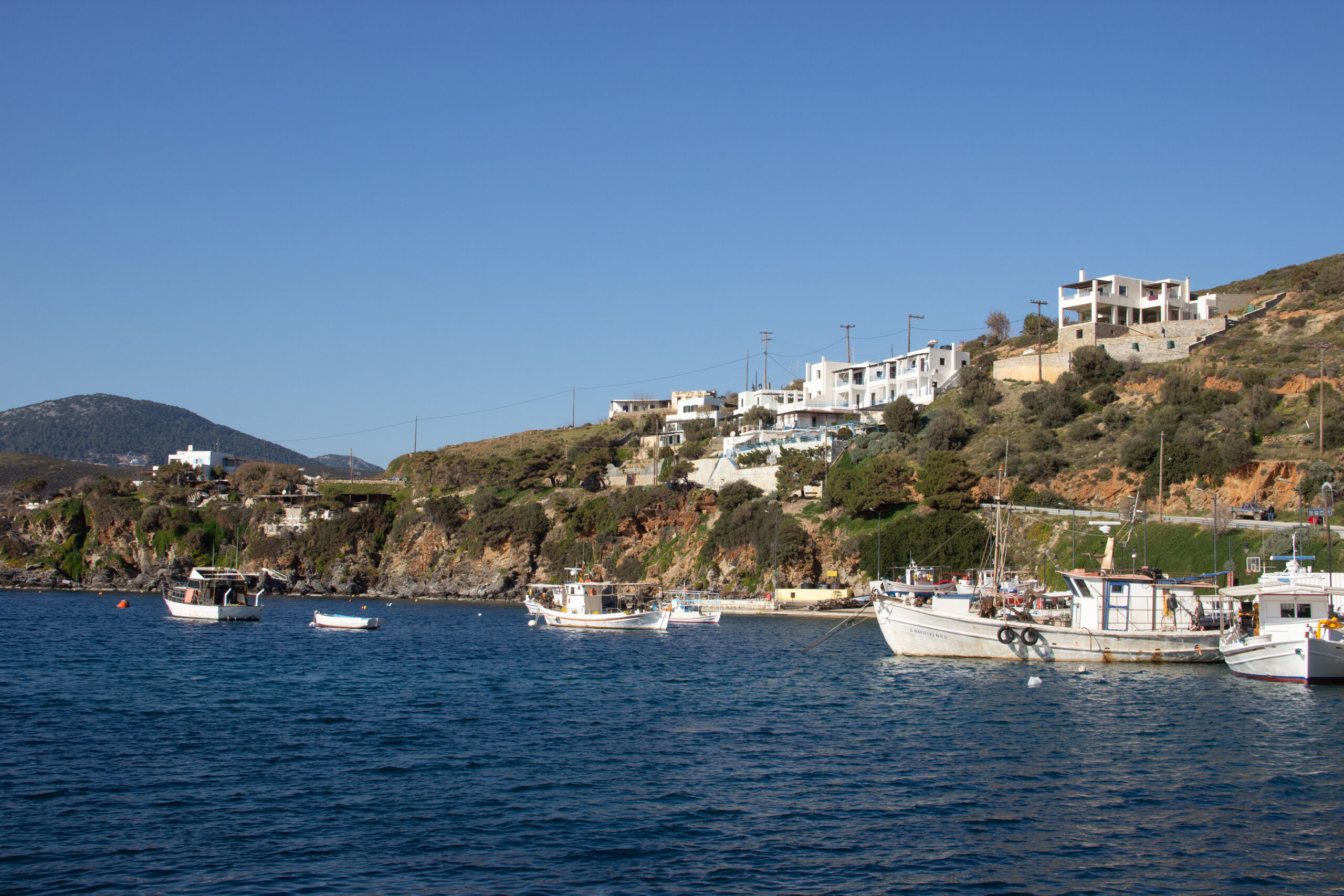 Scenic bay scene in Skyros with small boats docked in deep blue waters next to a low foliage covered and white-house set cliff.