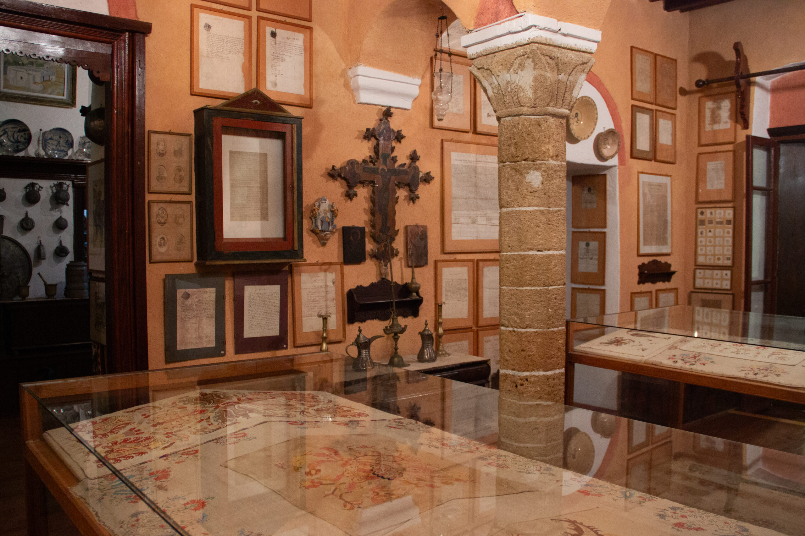 Interior of a room the Skyros Faltaits Museum with stone columns, woven fabric protected in glass cases and walls filled with framed scripts.